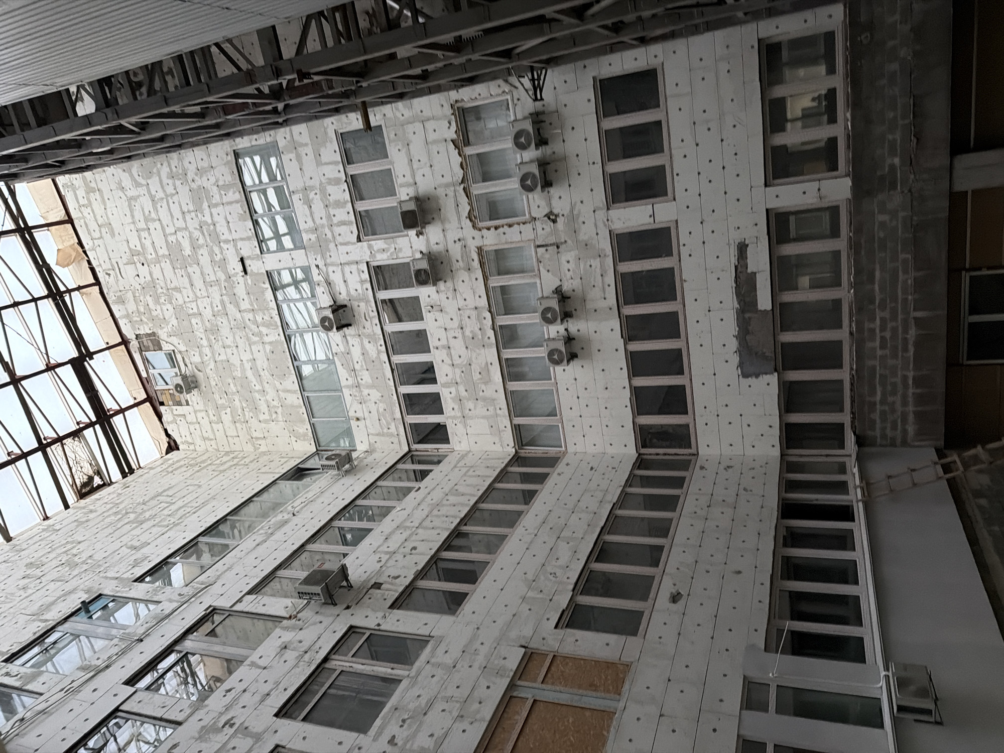 The image depicts an interior courtyard of a multi-story building, likely under construction or renovation, in Kyiv, Ukraine. The building's exterior walls are covered with a light-colored, textured material, possibly insulation panels, dotted with numerous small, dark fasteners or points. Multiple rows of windows, many with light-colored frames, punctuate the walls. Several windows are boarded up with light brown material. Air conditioning units are mounted on the exterior walls at various levels. A metal industrial structure, possibly a scaffolding or part of the building's framework, is visible on the right side of the frame. The roof appears to be a large, open skylight with visible metal support beams, some of which are damaged or incomplete. The ground level of the courtyard shows a brick wall and further windows. A wooden ladder leans against the interior wall near the bottom left. The overall impression is one of an unfinished or damaged urban construction site.