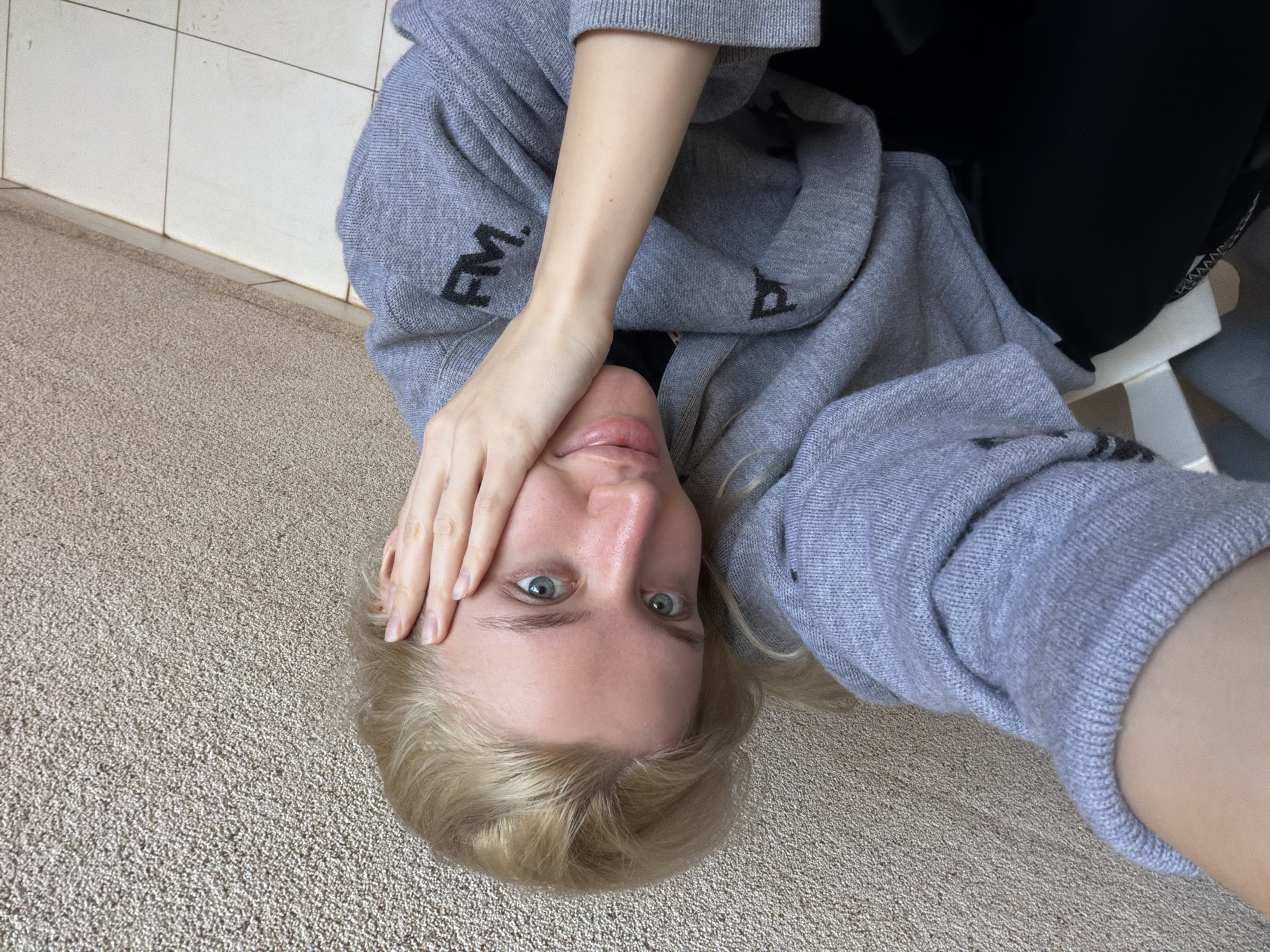 The image shows a person lying on their back on a carpeted floor. The person has blonde hair and light blue eyes. They are wearing a grey sweatshirt with black lettering. The person's right hand is resting on their forehead. There are white tiles and a baseboard visible in the background. The location in the city of Nerin, Montenegro, is unknown from this image.