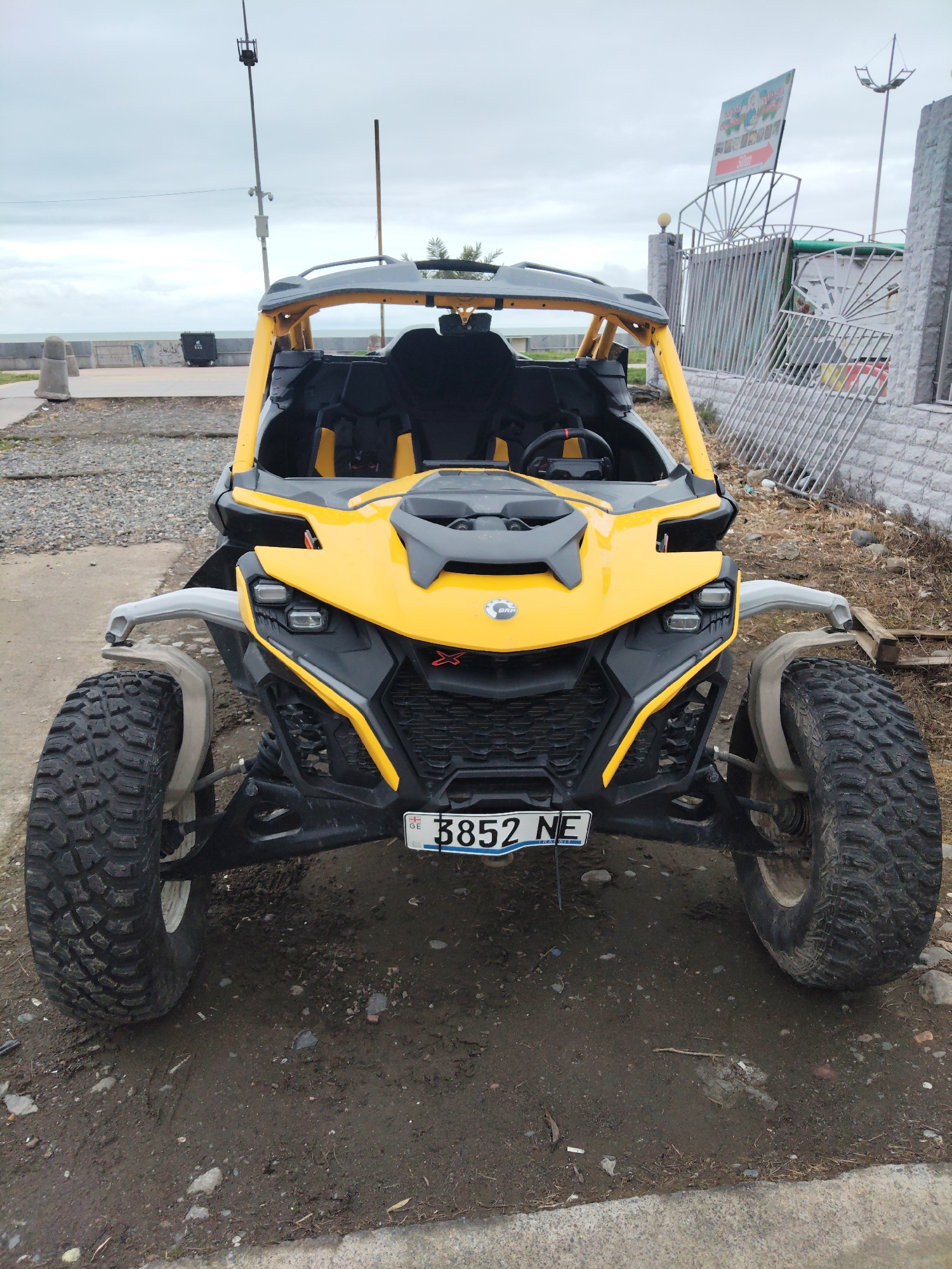 A yellow and black Can-Am Maverick X3 stands on a patch of muddy ground adjacent to a paved area, potentially a sidewalk. The vehicle has a Georgian license plate reading "GE 8852 NE". The background shows a coastal scene with a concrete barrier overlooking the sea, and a sign on a metal fence behind a stone wall. A tall mast with what appears to be a security camera is also visible in the distance. The sky is overcast, suggesting a cloudy day in Batumi, Georgia.