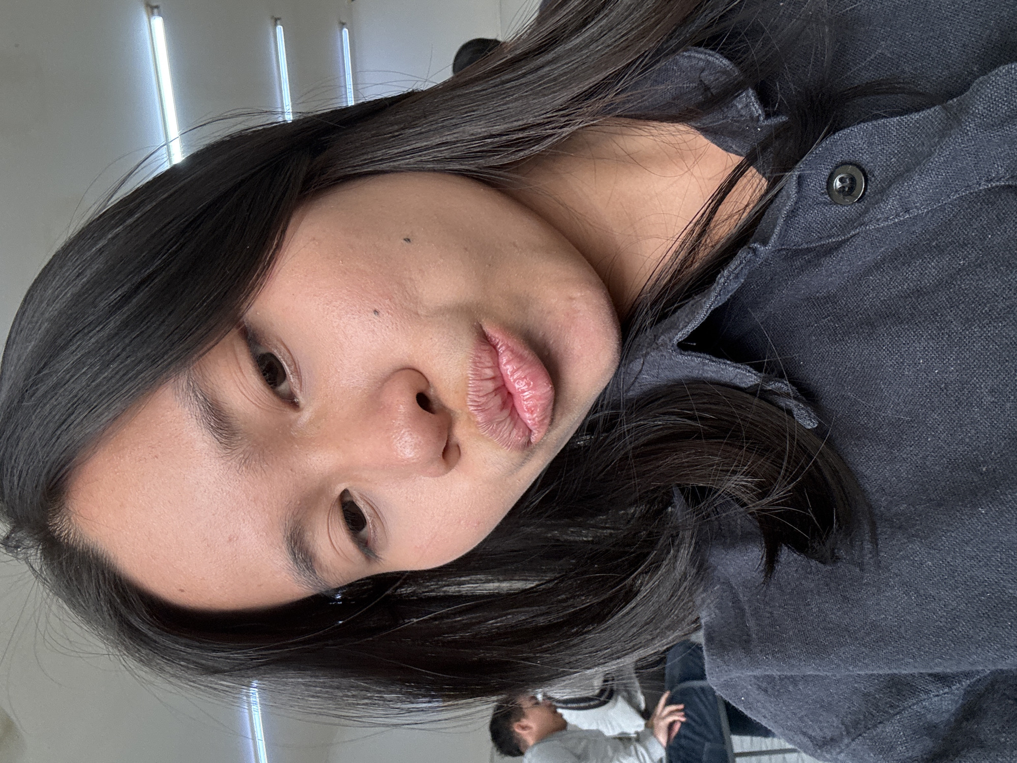 A young Indonesian woman of East Asian descent with long, dark hair is taking a selfie in what appears to be a classroom or common area. She is wearing a dark-colored collared shirt with a single black button visible at the neck. Her face is turned towards the camera, with her lips pursed in a playful "duck face" pose. She has dark eyes and a few subtle beauty marks on her right cheek.
In the background, out of focus, two other individuals are visible. One person, wearing a light gray long-sleeved shirt and dark pants, is seated on a dark surface, possibly a bench or couch, and appears to be looking down. Another person, further in the background and also out of focus, has dark hair and is partially visible. The environment is illuminated by fluorescent strip lighting fixtures on the ceiling, suggesting an indoor setting that might be a school or public building. There are no specific landmarks visible that would definitively place the location within East Jakarta, Indonesia, but the presence of individuals of East Asian descent is consistent with the region.