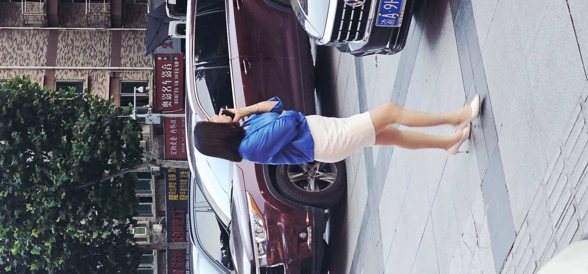 Here is a forensic description of the image:
**Scene Overview:** The image captures a street scene in what appears to be an urban environment. Buildings line the street, interspersed with trees.
**People:** The primary subject is a woman standing on the sidewalk. She is engaged in a phone conversation. She is wearing a blue blouse, a light-colored skirt, and white heels.
**Objects:** Several cars are parked along the sidewalk. Storefront signs written in Chinese characters are visible in the background.
**Actions:** The woman is talking on her phone. There is no other discernable activity captured in the static image.
**Environment:** The scene appears to be a sidewalk in a commercial district. The presence of cars and signs suggests a busy urban setting. Based on the signs present, the location is in Yuzhong District, China.