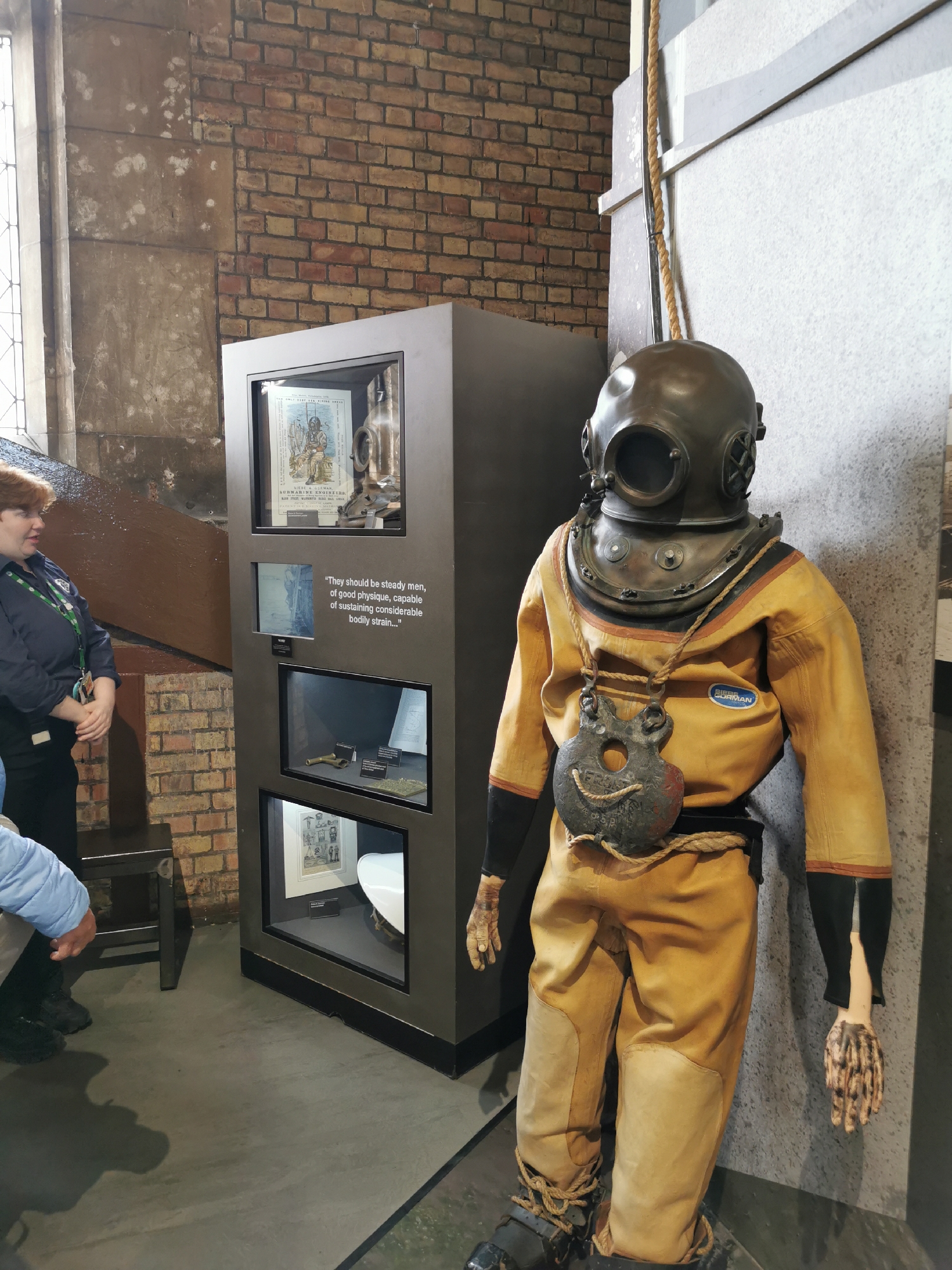 An indoor shot captures a vintage diving suit mannequin alongside a modern display case. The mannequin, positioned on the right, wears a heavy brass diving helmet, a yellowish-orange canvas diving suit, and appears to have lead weights around its waist and chest. A rope is tied around its waist and neck. Its hands, or gloves, are discolored and appear somewhat skeletal.
To the left of the mannequin is a tall, dark grey display case with multiple illuminated compartments. The top compartment holds what appears to be an old print or illustration depicting a diver, and some diving equipment. Below it, at eye level, a quote is displayed in white text against a dark background: "They should be steady men, of good physique, capable of sustaining considerable bodily strain..." Below this, another compartment shows smaller artifacts, possibly brass fittings or tools. The lowest visible compartment also contains an illustration and what looks like a white ceramic part of equipment.
A woman with reddish-brown hair, wearing a dark blue jacket with a green lanyard around her neck, stands to the left of the display case, looking towards it. Her expression is attentive. Part of another person is visible in the lower left, bent down near a dark wooden stool.
The background features an exposed brick wall on the left, transitioning to a lighter, possibly stone or concrete wall on the right. A large window with leaded panes is partially visible on the far left. The floor is dark grey and appears to be concrete or similar material. The scene suggests a museum or exhibition setting, showcasing historical diving equipment. Text visible on the diving suit's chest patch reads "BAMAN". Other partial text from the display cases includes "ONL" and "BMARINE ENG".