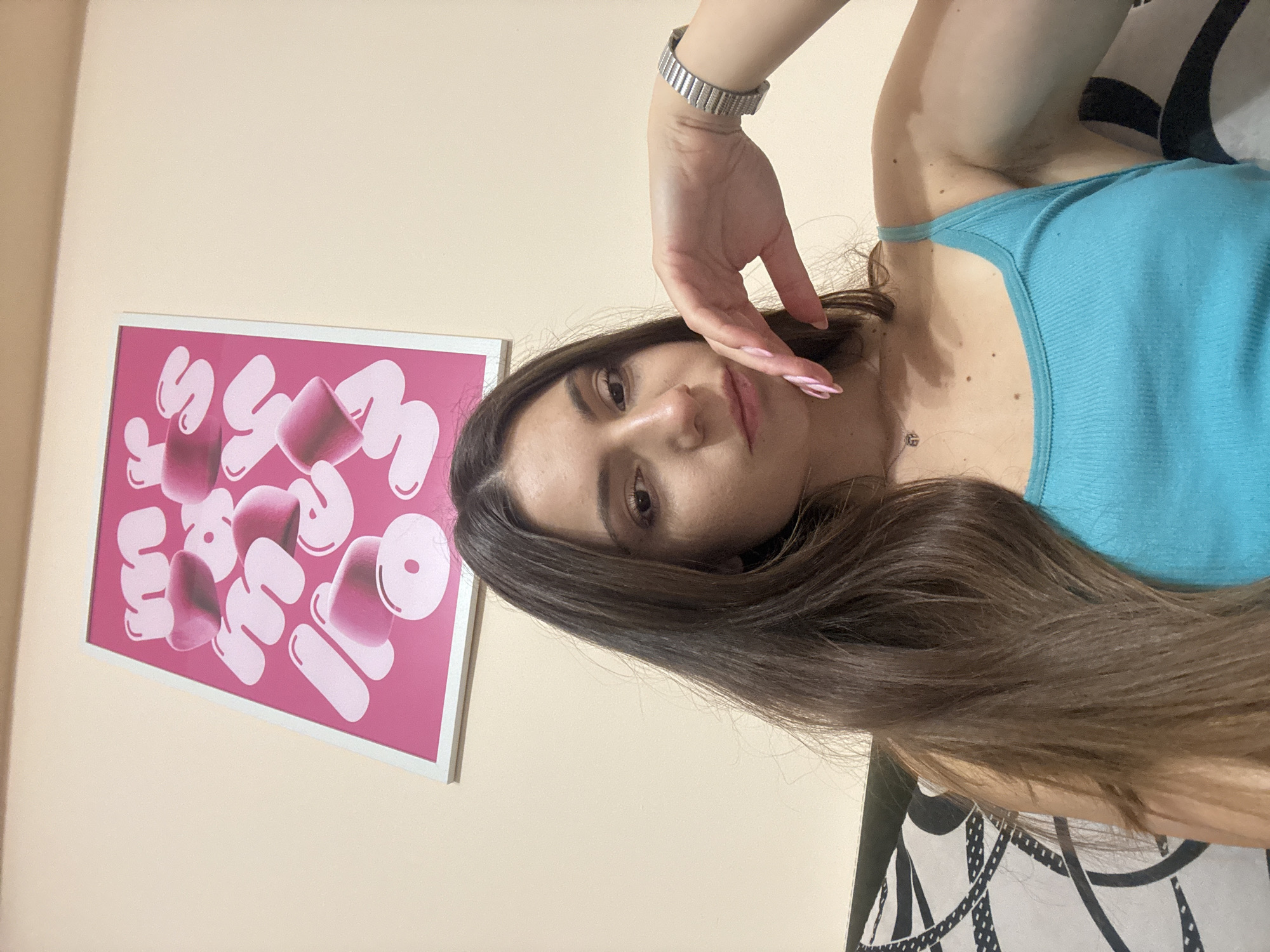 An indoor, eye-level, selfie photograph captures a young adult woman seated against a wall with a framed pink poster featuring white and pink block letters. The woman has long, dark brown hair and is wearing a blue tank top. Her right hand rests on her chin, and she is looking towards the camera. A metallic watch is visible on her left wrist. A pillow with a black and white patterned cover is partially visible to her left. The environment appears to be a domestic setting, possibly a bedroom or living room, with a plain, light-colored wall. The image is undated and undated, and its specific location within Varna, Bulgaria, cannot be determined from the visual information provided.