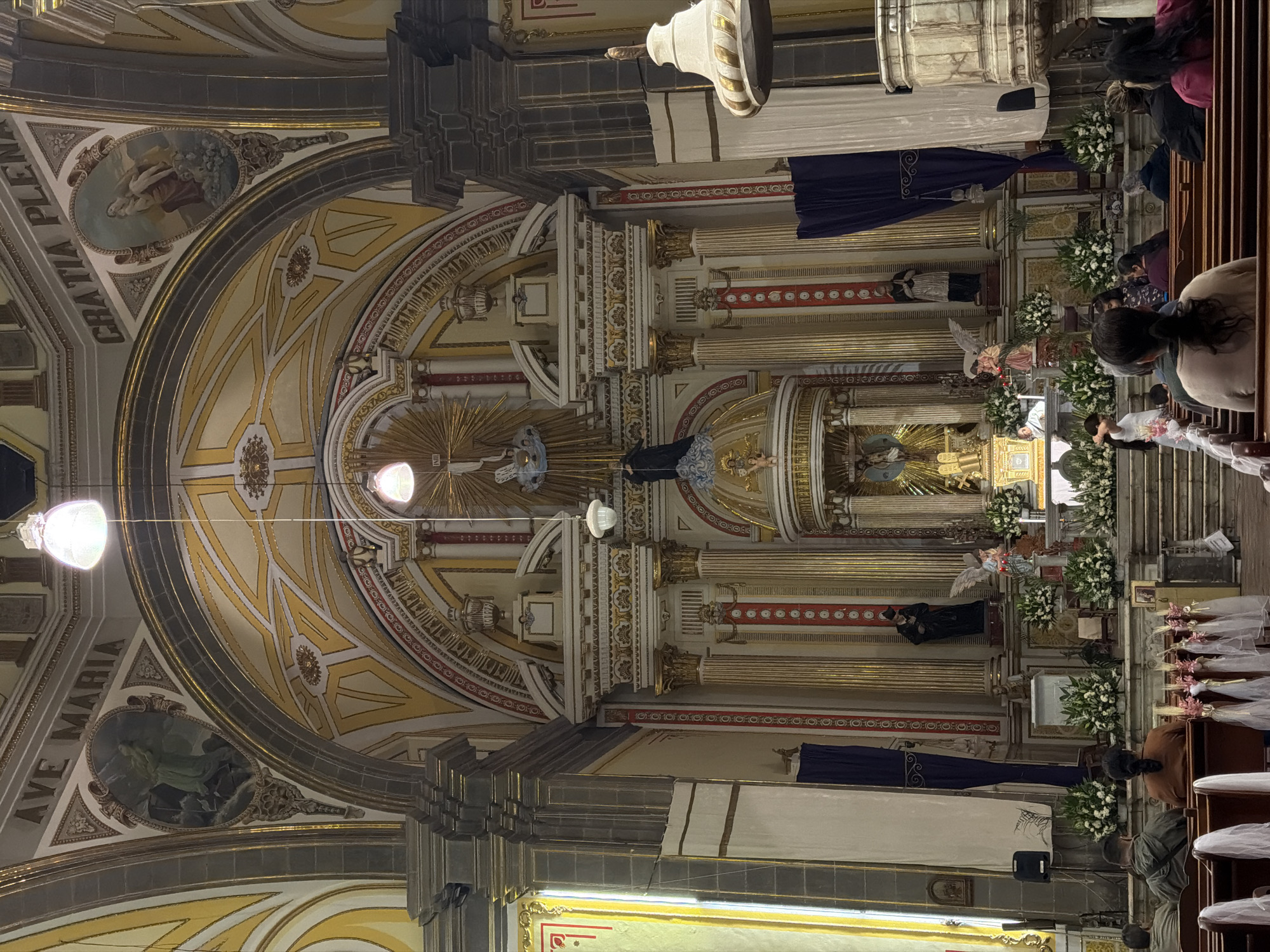 The image shows the interior of a church, with a view towards the altar. The building features a high, arched ceiling adorned with gold and white ornamentation. The ceiling paintings are of religious scenes. The altar area includes columns, statues, and floral arrangements. People are seated in pews, likely attending a service. The image likely depicts the Church of Santo Domingo in the city of Puebla, Mexico.