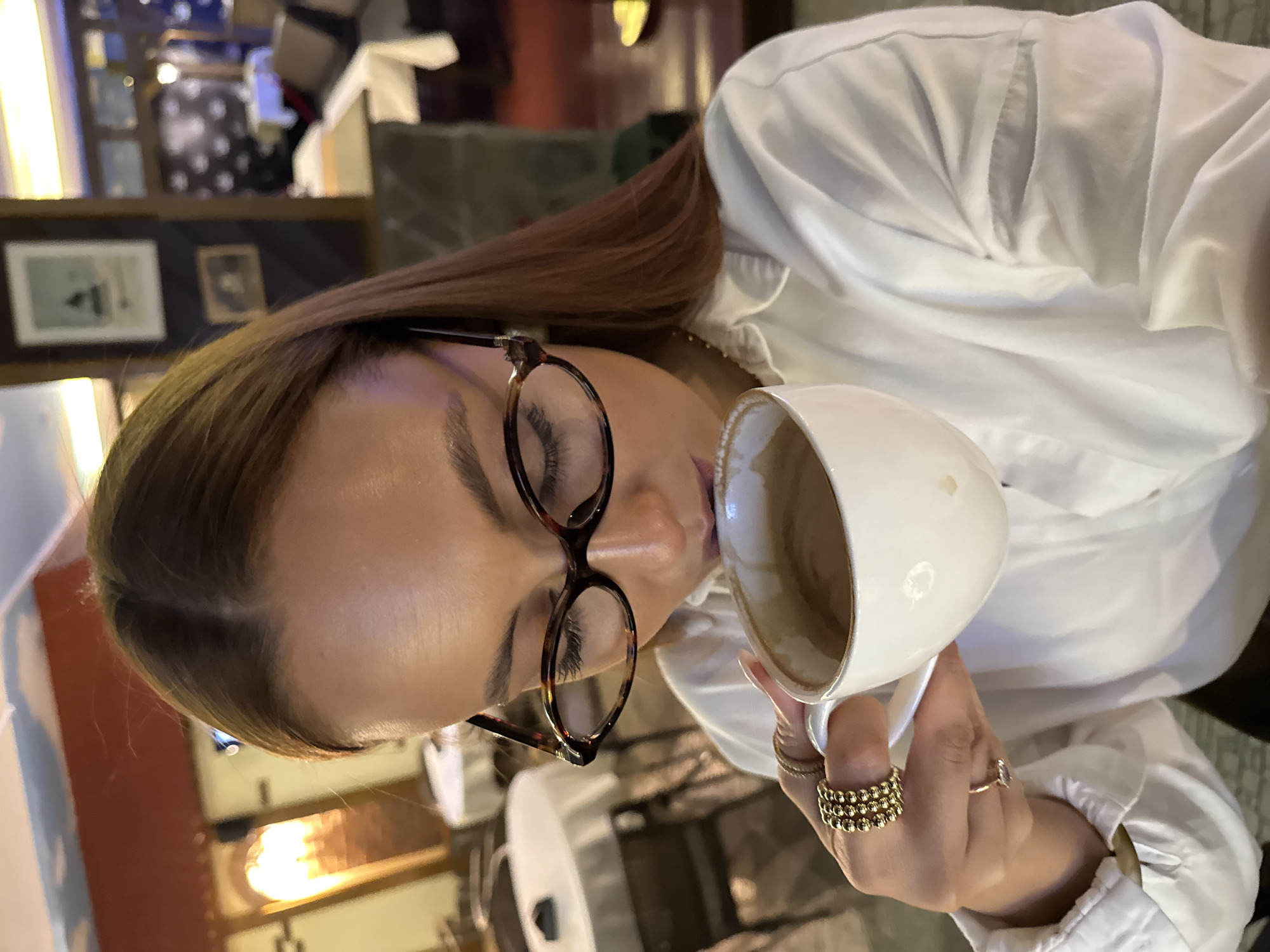 A medium close-up, horizontally oriented shot depicts a person, likely a woman, indoors, engaged in drinking from a white mug. She is positioned on the left side of the frame, facing right, with her head tilted slightly downwards towards the cup. Her eyes are closed or looking down, concentrating on the beverage. She has light to medium brown hair, parted on the side, and wears stylish round, tortoise-shell patterned eyeglasses. Her eyelashes appear long.
She is dressed in a white long-sleeved shirt with a collar and some visible pleating or ruffles near the neckline. Her left hand holds the white mug, which contains a light brown beverage, possibly coffee or a latte, with some residue visible on the inside. On her left hand, she wears several rings: a prominent multi-row ring composed of small golden beads on her ring finger, and at least two other delicate golden rings on adjacent fingers. Her fingernails are manicured.
The background is dimly lit and appears to be an interior space, possibly a café or restaurant. On the upper left, blurred elements suggest shelves or displays with items like framed pictures and possibly small decorative objects. A rectangular framed picture with a faint white border and dark content is visible on a dark wall or shelf. Soft, warm lighting sources are scattered in the background, creating bokeh effects. The overall atmosphere seems relaxed and intimate. There is no visible text in the image. The location is contextualized as Chaikovsky, Russia, though no specific visual cues for this city are present.