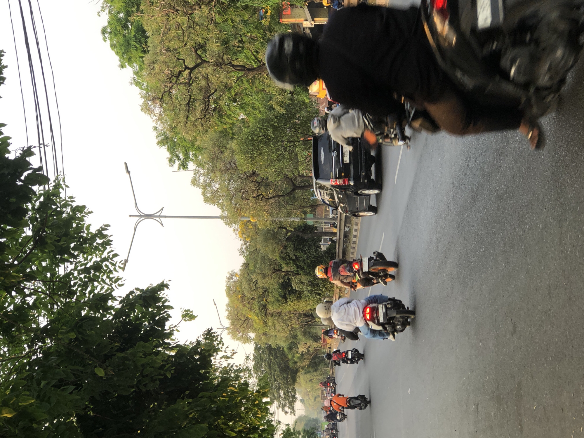 The image depicts a street scene in Kludan, Indonesia, characterized by a moderate flow of traffic. In the foreground, a person on a black motorcycle wearing a black helmet and black long-sleeved shirt is riding on the right side of the road, partially obscuring the view of the motorcycle behind. Several other individuals on motorcycles are visible in the middle ground, traveling in the same direction. One person on a white motorcycle with blue jeans is in the center, while another on a motorcycle with an orange rear wheel and a red and yellow jacket is further ahead. A black SUV is also present in the traffic. The environment is an urban road lined with trees, with a clear sky and streetlights visible. The image captures a typical moment of daily commute in a Southeast Asian city.