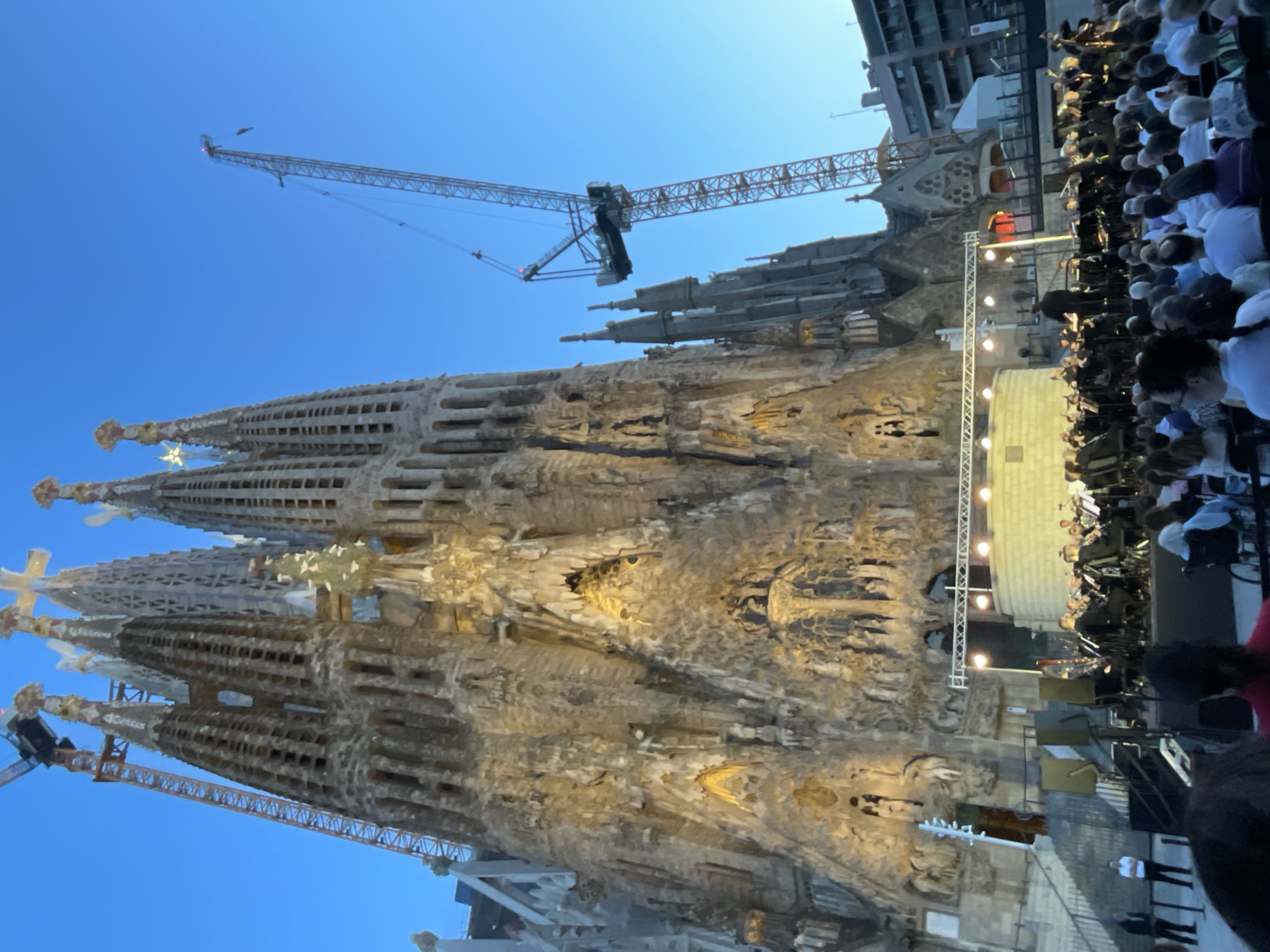 The image depicts an outdoor evening concert in front of the Sagrada Familia basilica in Barcelona, Spain. A large orchestra is performing on a stage set up on the grounds of the basilica, illuminated by stage lights. Spectators are seated in front of the stage, some in wheelchairs. The basilica, a UNESCO World Heritage Site, is prominently featured in the background, with its intricate facades illuminated and scaffolding and cranes visible, indicating ongoing construction. The sky is a clear blue, suggesting it is twilight. The overall scene captures a cultural event taking place at a significant architectural landmark.