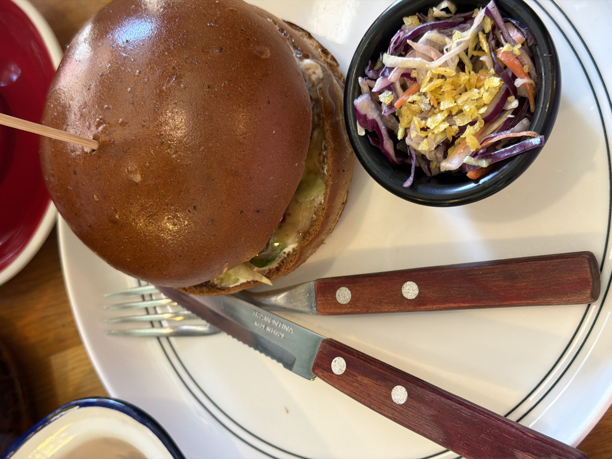 A close-up, high-angle shot captures a food presentation on a white plate with a double black line around its rim. The main element is a large, shiny, dark brown burger impaled with a toothpick. To the right is a small, black bowl filled with red cabbage slaw, shredded carrots, and a crispy topping. Below the burger, a silver fork and knife with wooden handles are placed horizontally on the plate. The knife's blade is serrated and marked "TRAMONTINA, MADE IN BRAZIL." A red bowl is partially visible on the upper left, and a blue-rimmed dish is partially visible on the lower left. The food is positioned on a wooden surface. The location is Saint Petersburg, Russia.