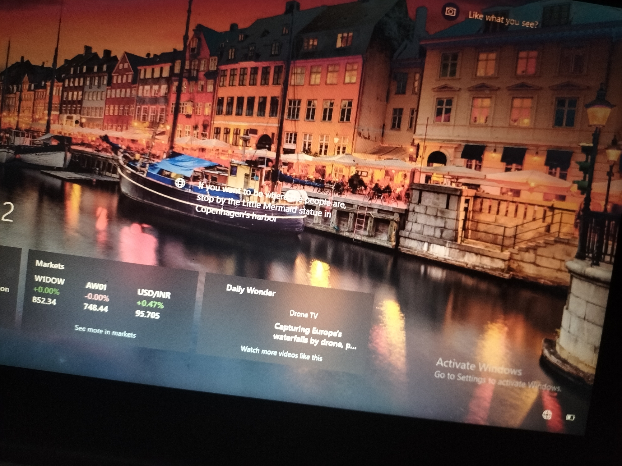 The image depicts a computer screen displaying a lock screen or desktop wallpaper. The background features a scenic view of a canal lined with colorful buildings, likely in a European city. Boats are docked along the canal. Overlaying this background are various informational widgets and text elements.
On the left side, a section labeled "Markets" displays stock market data including "WIDOW" with a value of "+0.00%" and "852.34", and "AW01" with "-0.00%" and "748.44". Below this, "USD/INR" shows "+0.47%" and "95.705". A text prompt "See more in markets" is also present.
Another widget, labeled "Daily Wonder", presents "Drone TV" with the text "Capturing Europe's waterfalls by drone, P...". A call to action, "Watch more videos like this", is also visible.
In the upper right corner, text reads "Like what you see?".
Towards the bottom right, the text "Activate Windows" is displayed, followed by "Go to Settings to activate Windows.".
The image does not contain any elements that suggest the location is Jalingo, Nigeria. The background imagery and the text content (e.g., "Copenhagen's harbor", "Drone TV", "Europe's waterfalls") indicate a European setting and a digital interface rather than a specific geographical location in Nigeria.