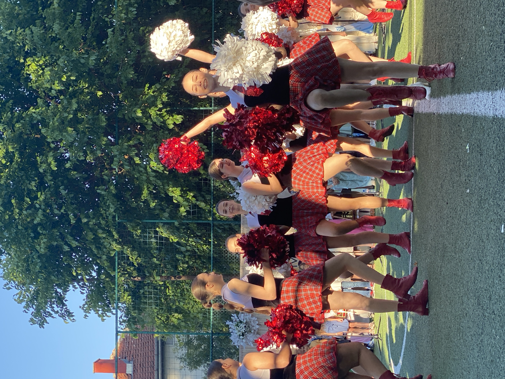 The image shows a group of young women in cheerleader outfits, performing on a green turf field. They are wearing red and white pom-poms and plaid skirts. The women are in various poses, with some holding their pom-poms up in the air. In the background, there are trees, a fence, and buildings. The scene appears to be outdoors, possibly at a sporting event or a school function. The image does not provide specific details to pinpoint the location in the city of Pleven, Bulgaria.