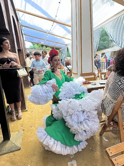 The scene depicts a woman in a traditional flamenco dress, with layers of white ruffles on the sleeves and skirt over a green base, seated at an outdoor table. She has a red floral arrangement in her hair, earrings, and wears a green dress with a low neckline. The woman appears to be smiling and looking towards the camera.
She is sitting at a wooden table in what seems to be a tent or temporary structure with a white and gold border trim around the columns. The tent is covered with a white canopy and decorative lights. In the background, other people are visible, including a woman in a black dress holding a purse and another woman with dark curly hair wearing a crocheted shawl over her shoulders.
The ground appears to be dusty and sandy, with scattered debris. The scene conveys a festive or celebratory atmosphere. The image was taken in Jerez de la Frontera, Spain, specifically during the Feria del Caballo, a local horse fair known for its traditional attire and festivities.
