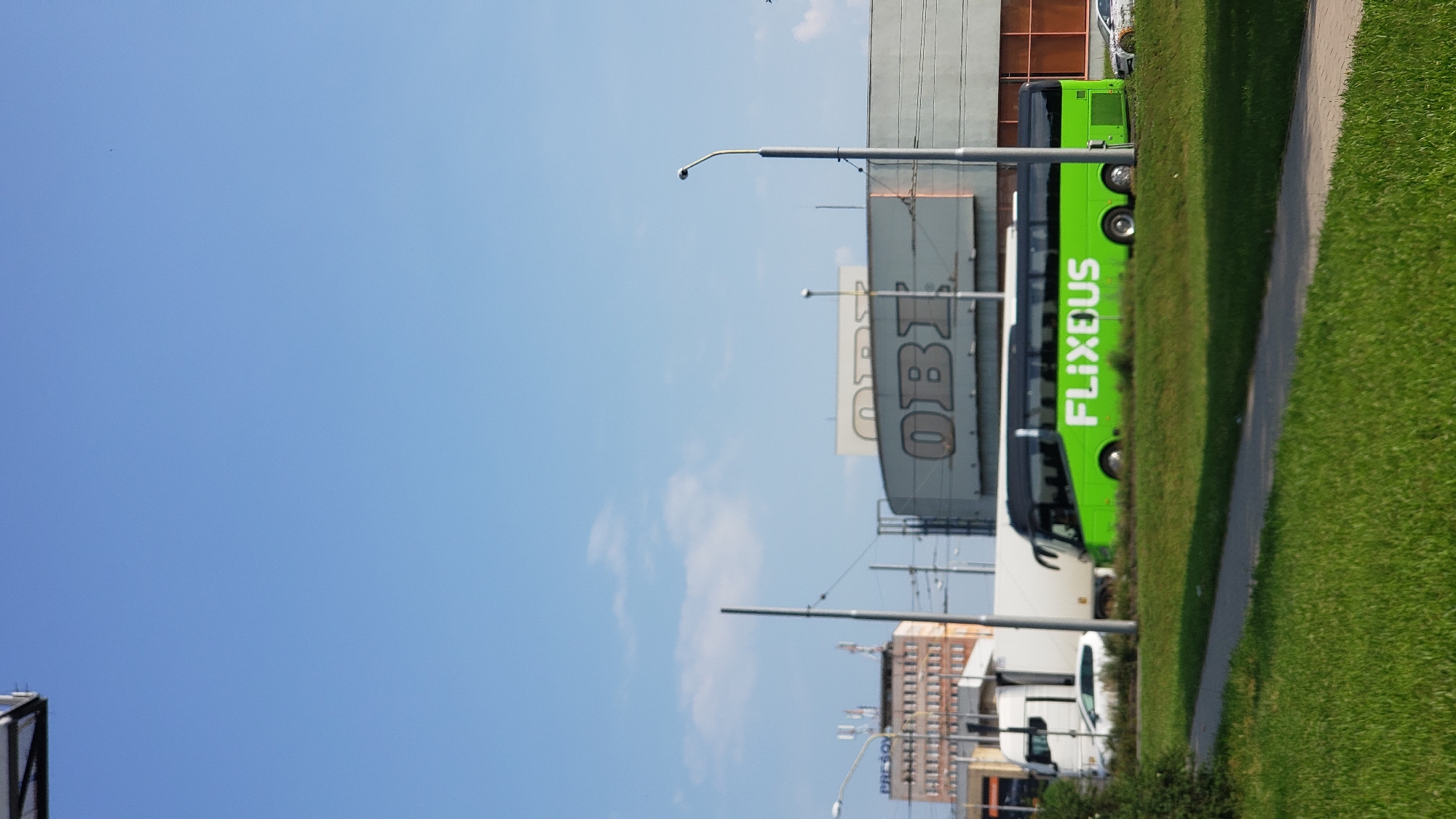The image captures a bright, sunny day in the city of Prešov, Slovakia, with a clear blue sky and scattered clouds. In the foreground, a neatly trimmed green lawn is visible, with a paved pathway running through it. Dominating the mid-ground is a bright green FlixBus, its logo prominently displayed on its side, traveling along a road. Behind the bus, a white truck and a white car are also present, suggesting moderate traffic. Further in the background, a large building with "OBI" signage is visible, indicating a commercial area. The overall scene portrays a typical urban landscape on a pleasant day.