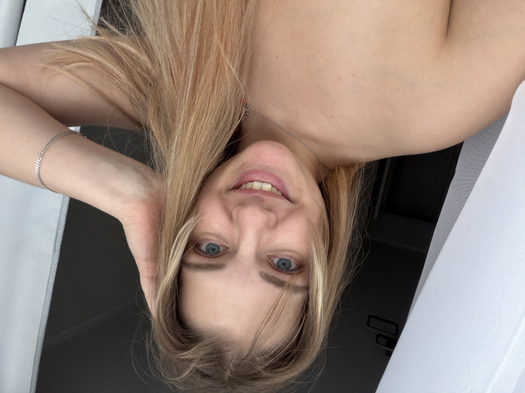 A woman with blonde hair is seen in an upside-down close-up. She has blue eyes and a slight smile. She is wearing a silver bracelet on her left wrist and a delicate necklace. She is holding her head with one hand. The background appears to be a window with white curtains and a dark interior. There is no information in the image to determine the location as Kazan, Russia.