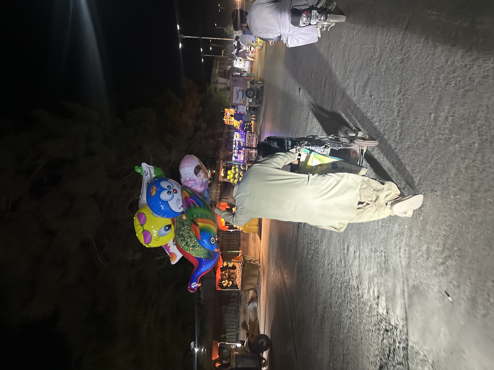 A vendor on a bicycle is positioned on a street in Quetta City Tehsil, Pakistan. He is selling an assortment of colorful character balloons. The street is illuminated by streetlights and traffic. Other people are also riding motorbikes.