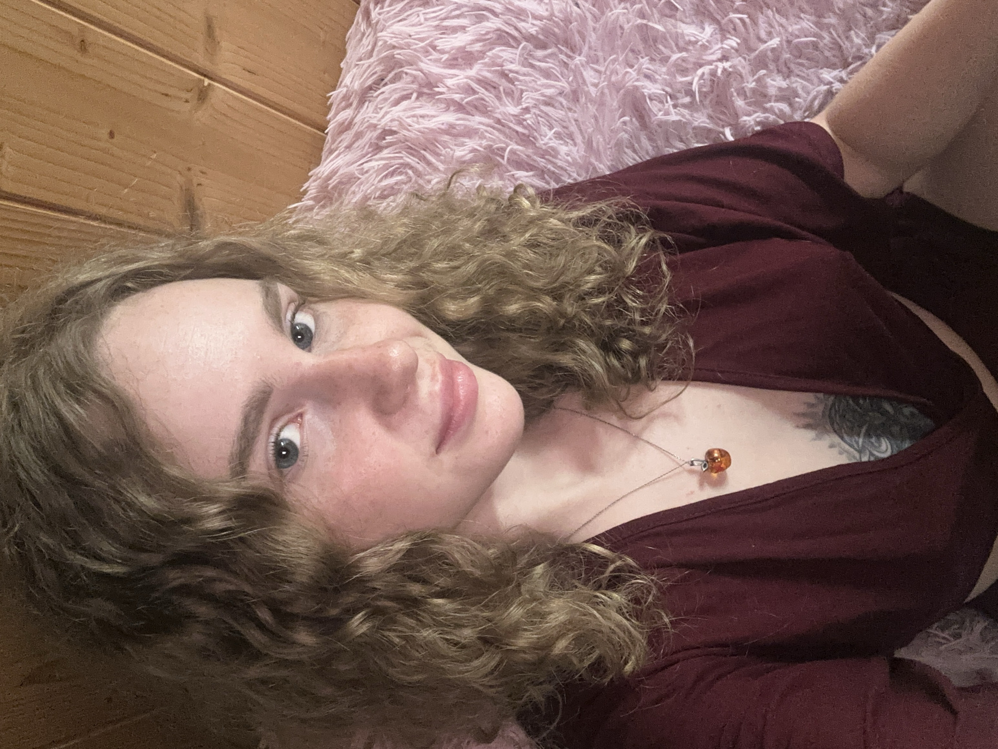 A person with fair skin and curly, light brown hair is lying down. They are wearing a maroon top and a necklace with an amber-colored pendant. A tattoo is visible on their chest. The person is lying on a pink, shaggy blanket with a light brown wooden surface visible in the background. The location is not possible to determine from this image.