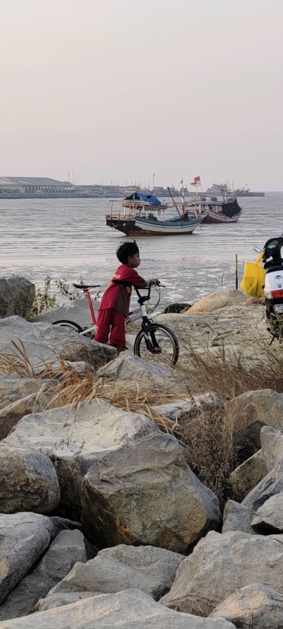 A young child wearing a red shirt and pants stands next to a bicycle on a rocky shoreline. The child is facing right, looking out at a body of water where several boats are docked. The water appears calm and reflects the overcast sky. In the background, there is a long building with a row of lights, possibly a pier or a waterfront structure. The foreground is dominated by large, grey rocks of varying sizes, with some dry grass growing between them. To the right of the frame, a portion of a white motorcycle with a yellow bag is visible. The scene suggests a quiet, outdoor setting near a harbor or coastal area in Tasikagung, Indonesia.