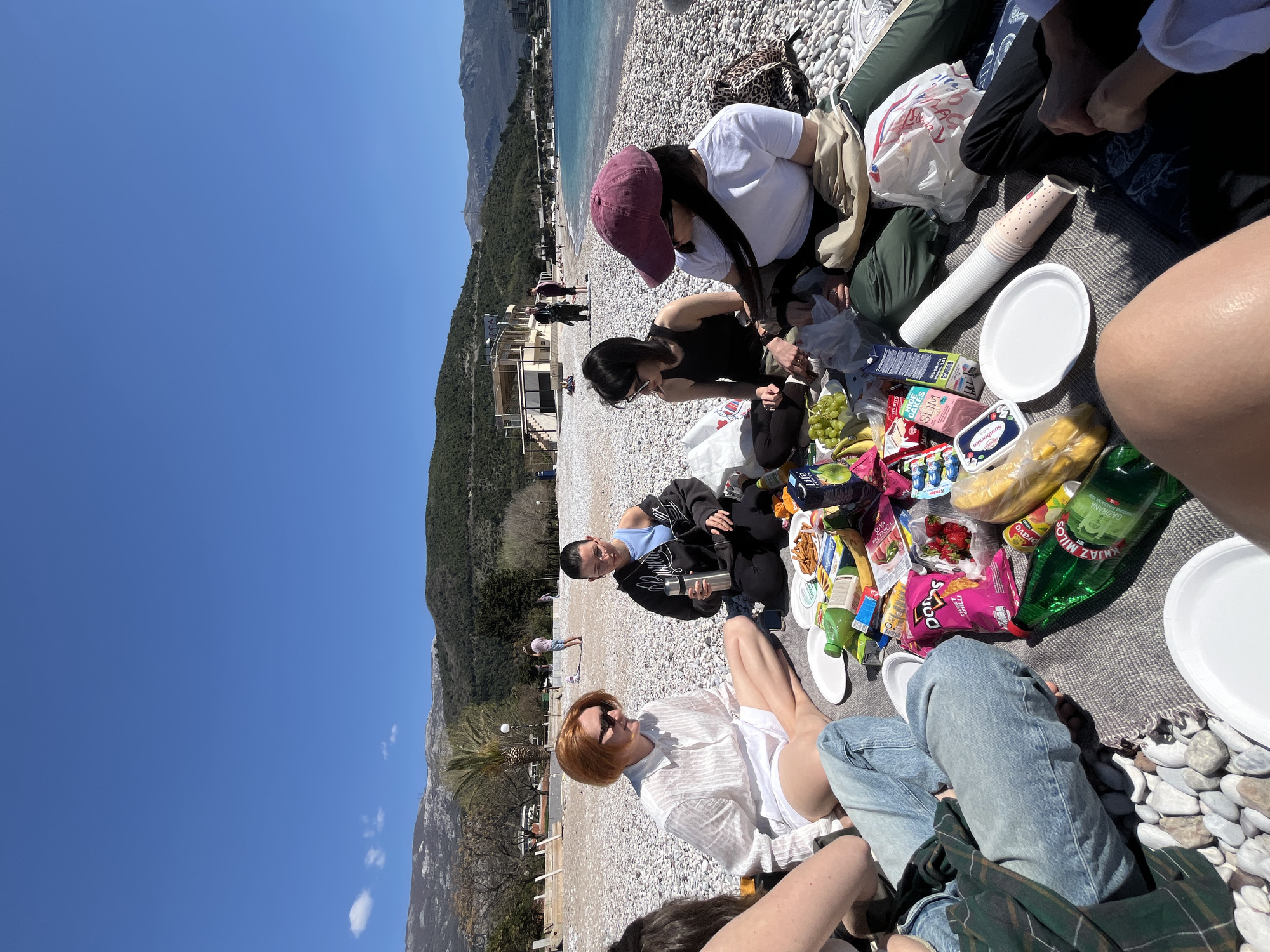 A group of people are having a picnic on a pebble beach. The scene includes five individuals, with focus on a picnic spread of food and drinks. The setting is the Mogren Beach in Budva, Montenegro, recognizable by the distinct landscape of rocky hills meeting the Adriatic Sea.
