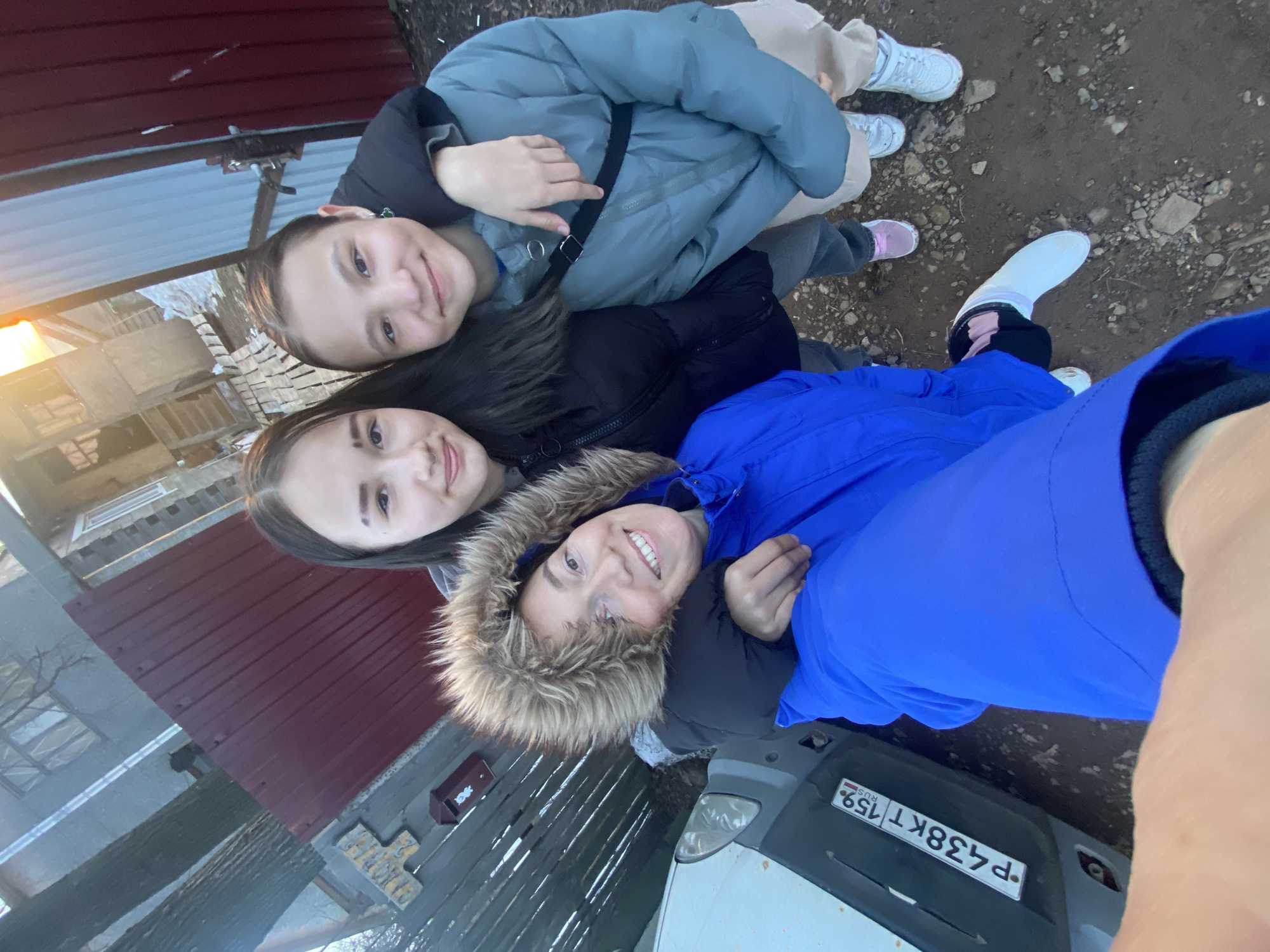 Three smiling females are captured in a selfie-style photograph outdoors in Ольховка, Russia, during what appears to be late afternoon or early evening. The person holding the camera, an adult female with light-colored hair, is positioned on the left, wearing a bright blue hooded jacket with a fur-lined hood. Her right arm is around the shoulder of a younger female in a black padded jacket, who is positioned centrally. The central female's left arm is around the shoulder of another younger female on the right, who wears a light blue/grey padded jacket, grey pants, and light-colored sneakers. All three individuals are smiling directly at the camera.
The background features a partial view of a white vehicle on the far left, displaying a Russian license plate "P438KT 159 RUS". Behind the subjects, a dark red corrugated metal fence or gate spans across the upper left and center. To the left of this fence, a wooden slatted fence is visible, bearing a small reddish-brown mailbox and a wooden sign partially reading "САДОВАЯ 22". Beyond the fences, parts of a grey building with windows and another wooden structure with steps are discernible. Two tree trunks are visible on the far left. The ground is composed of uneven dirt and small rocks. A warm, low sun, suggesting dusk or dawn, is visible in the upper right, casting a golden glow.