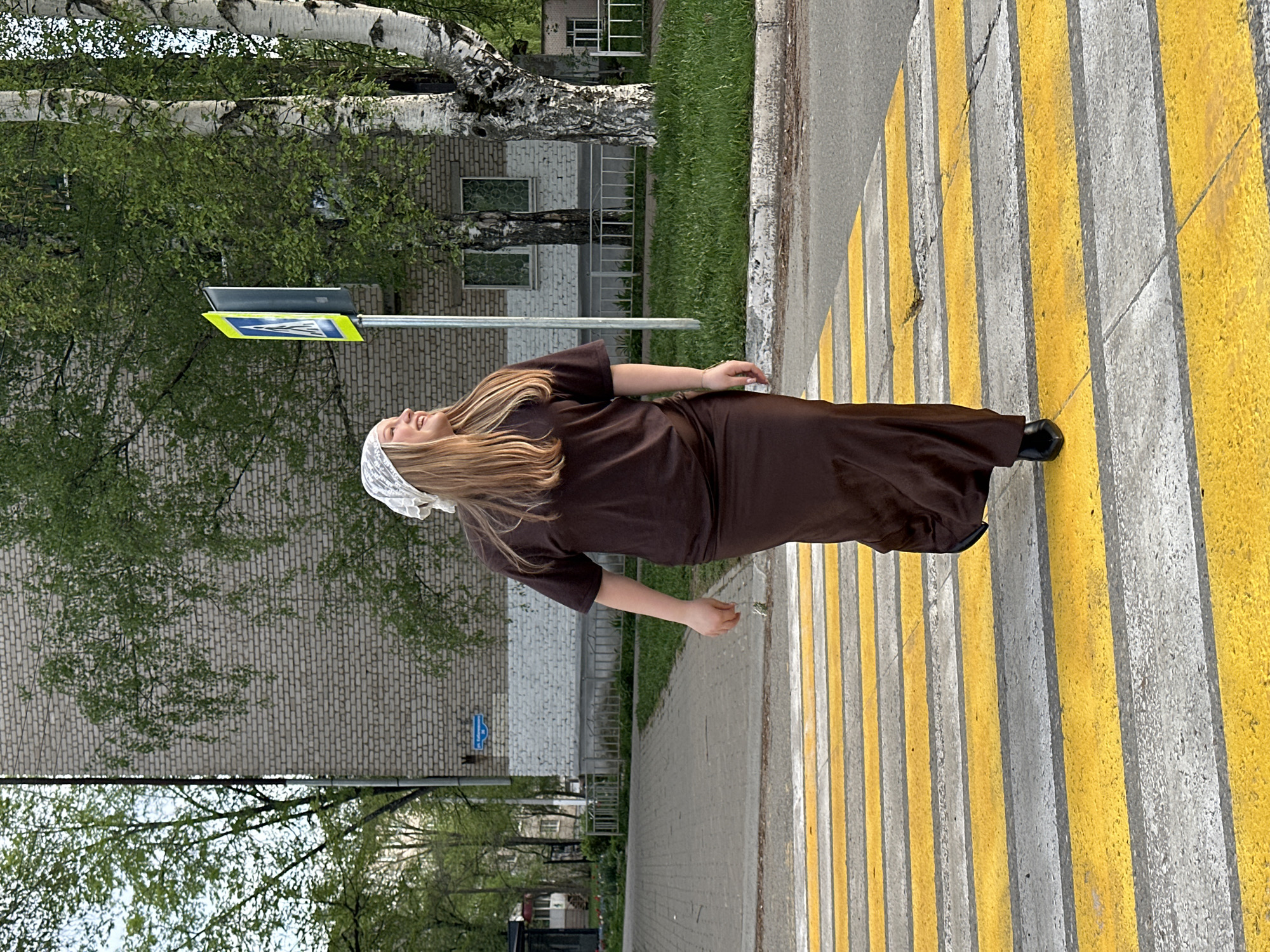 A photograph taken in the city of Chaikovsky, Russia, depicts a woman on a crosswalk. The crosswalk is painted with alternating white and yellow stripes. The woman is wearing a brown dress, black shoes, and a white headscarf. She is lying on her back on the crosswalk, with her arms extended to the sides and her head tilted back. A pedestrian crossing sign is visible behind her, along with a brick building and trees.