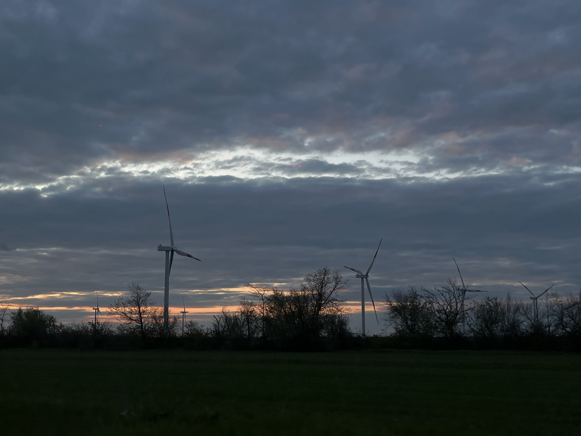 The image shows a landscape with wind turbines at dusk. A field of dark green grass occupies the foreground. A line of trees and bushes stretches across the middle ground, behind which several wind turbines are visible. The sky is filled with layers of gray and dark clouds, with a sliver of light appearing near the horizon. A faint, reddish object is visible in the upper left quadrant of the sky. It's impossible to verify from the image alone that the location is Port-Katon, Russia.