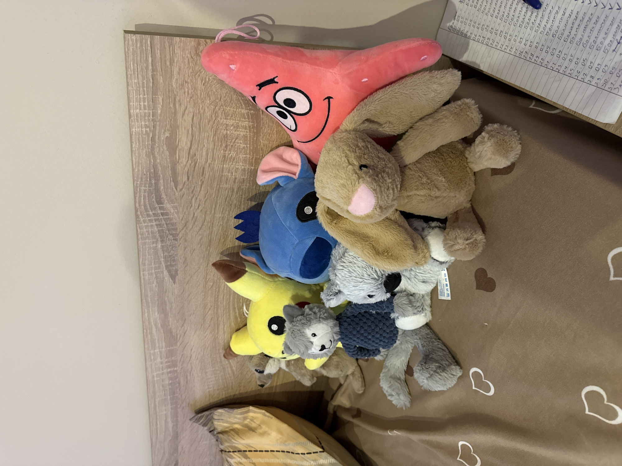 A stack of stuffed animals, including a pink Patrick Star, a brown bunny, a blue Stitch character, a yellow Pikachu, and a gray koala, lies on a bed with brown bedding adorned with white heart patterns. Next to the stack is a notebook filled with handwritten Bulgarian text, with some entries marked with a checkmark. The background consists of a wooden surface and a plain white wall. There is no information to determine the location as being in Lovech, Bulgaria.