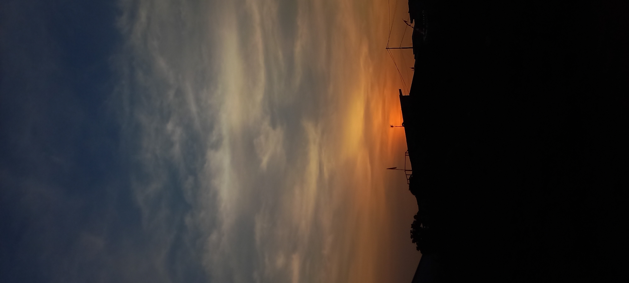 The image depicts a silhouetted landscape at dusk, likely in Loa Lepu, Indonesia, given the context. The sky is a gradient of deep blue at the top, transitioning through shades of grey and orange as it approaches the horizon. Wispy clouds are scattered across the sky, catching the last rays of sunlight.
In the foreground, the silhouettes of rooftops dominate the lower portion of the image. These appear to be residential or commercial structures. One prominent roof has a small flag on a pole, adding a touch of detail to the otherwise dark shapes. On the right side, utility poles and wires are also visible in silhouette against the sky. The overall impression is one of a quiet evening in a populated area as day turns into night. There are no visible people or specific actions taking place. The environment suggests an urban or semi-urban setting.