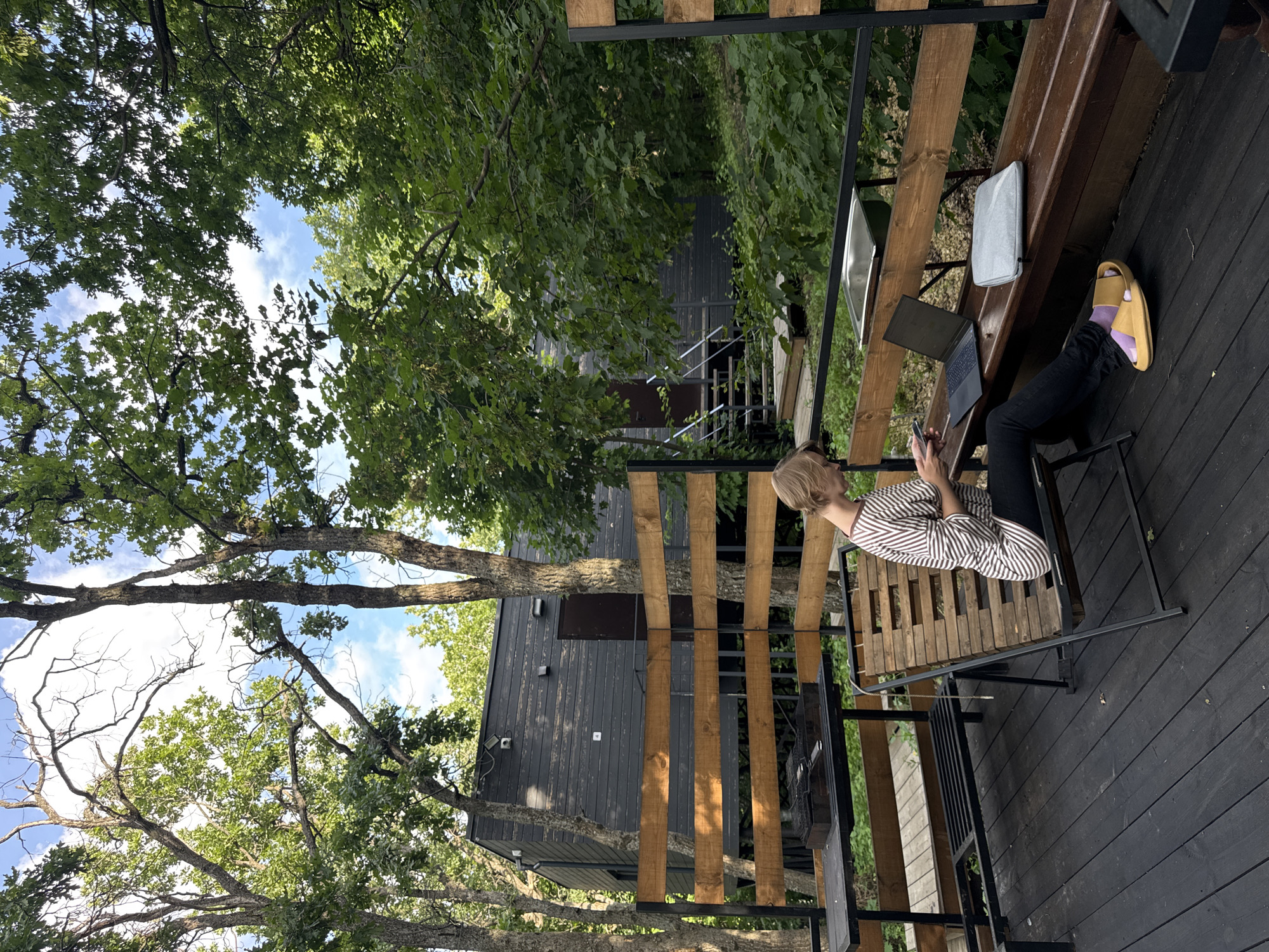 In a wooded outdoor setting in Ust-Kurdyum, Russia, a woman is seated on a wooden lounge chair on a dark wooden deck. She is wearing a striped long-sleeved shirt and black pants, with yellow slides on her feet. The woman is looking down at her phone, which she holds in her hands. A silver laptop is open on the deck in front of her, and a gray laptop sleeve is next to it on a wooden bench. Behind the bench, there is a metal sink. The deck overlooks a natural landscape with trees and a dark-colored building with a wooden fence in the foreground. The building appears to be a modern cabin or house constructed with horizontal wooden planks. The surrounding vegetation is lush and green, indicating a temperate climate.