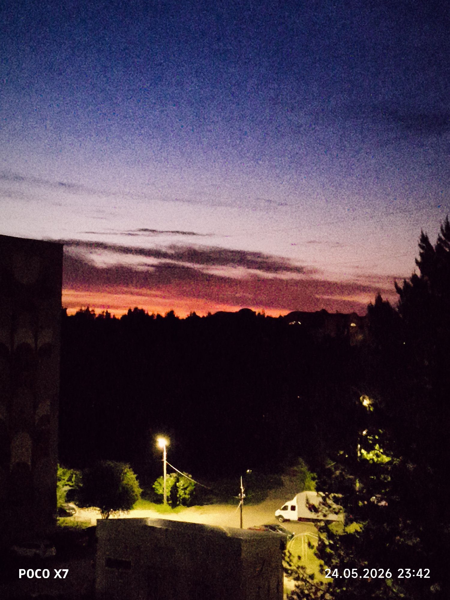 Captured from an elevated perspective, the image presents an outdoor scene at dusk or night in what appears to be a residential area. The sky transitions from a deep blue overhead to a band of pink and orange hues along the horizon, suggesting either sunset or sunrise. A dark silhouette of trees stretches across the horizon.
In the foreground, a section of a building's exterior is visible on the left, and a dense area of trees and bushes occupies the right side of the frame. Two streetlights illuminate the scene, casting a yellow glow on the surrounding foliage and structures. A white van is parked on a road or driveway. The overall environment seems tranquil and subdued.
The location is specified as Unknown, Unknown.