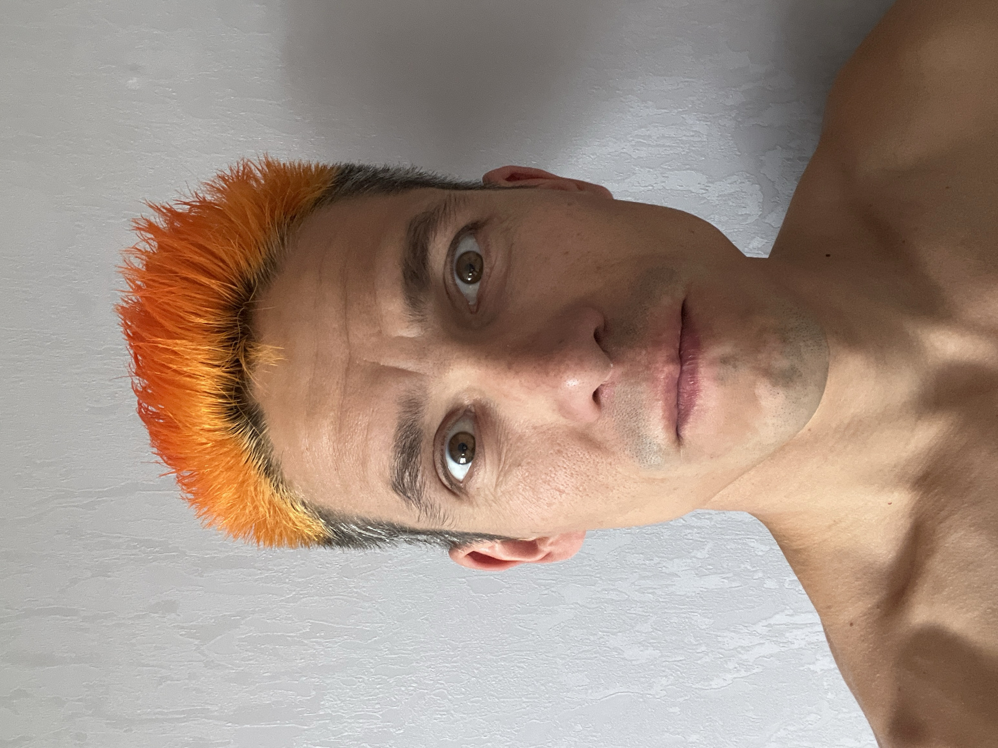 A close-up portrait features an adult individual looking directly at the camera. The person has short, spiky hair with vibrant orange and yellow-orange tips that transition to dark roots. They have brown eyes, dark eyebrows, and visible stubble on their face. Their expression is neutral, possibly slightly serious or contemplative. The person is shirtless, revealing their shoulders and upper chest.
The background is a plain, light-colored wall with a subtle, textured surface, suggesting an indoor setting. There are no distinct objects or discernible text in the image. The lighting appears even, illuminating the subject without harsh shadows. The image captures a moment, likely a self-portrait, in what could be a personal space within Chaikovsky, Russia.