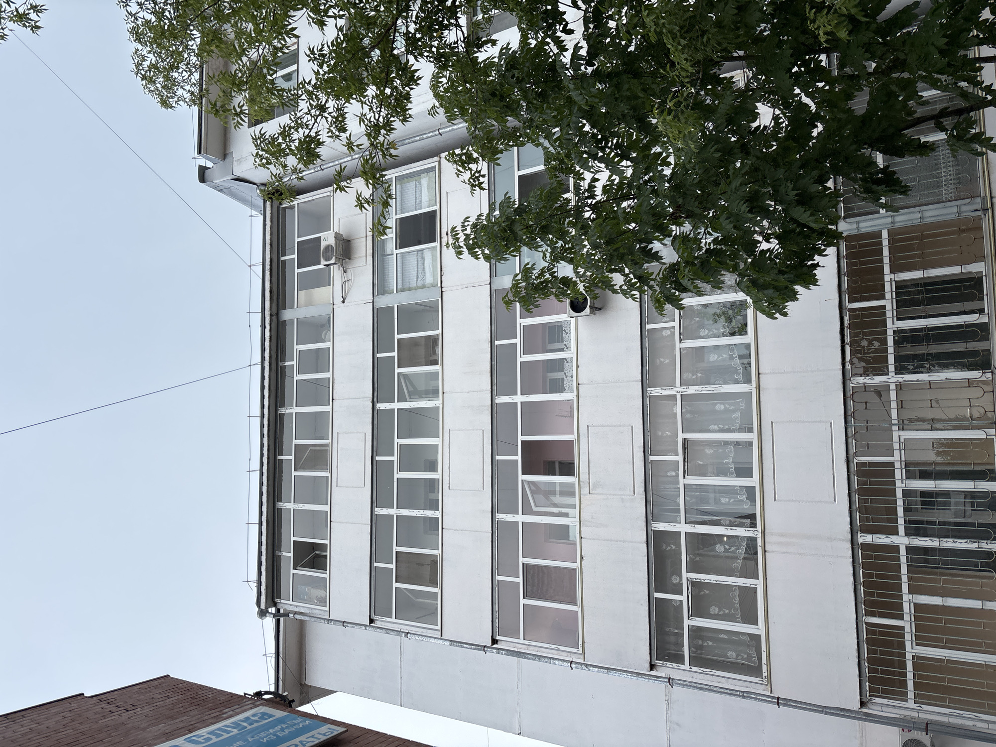 An exterior low-angle shot captures the side of a light-colored apartment building in Blagoveshchensk, Russia, against an overcast sky. The building features multiple levels of balconies, many with enclosed windows and some with open sections. Several air conditioning units are visible mounted on the exterior walls. A large green tree with dense foliage partially obscures the right side of the building. On the left, a portion of a red brick building with a blue sign is visible. The building appears to be a residential structure, likely an apartment block, typical of urban environments. The overall scene is mundane and devoid of human activity, focusing on the architectural details and the natural element of the tree.