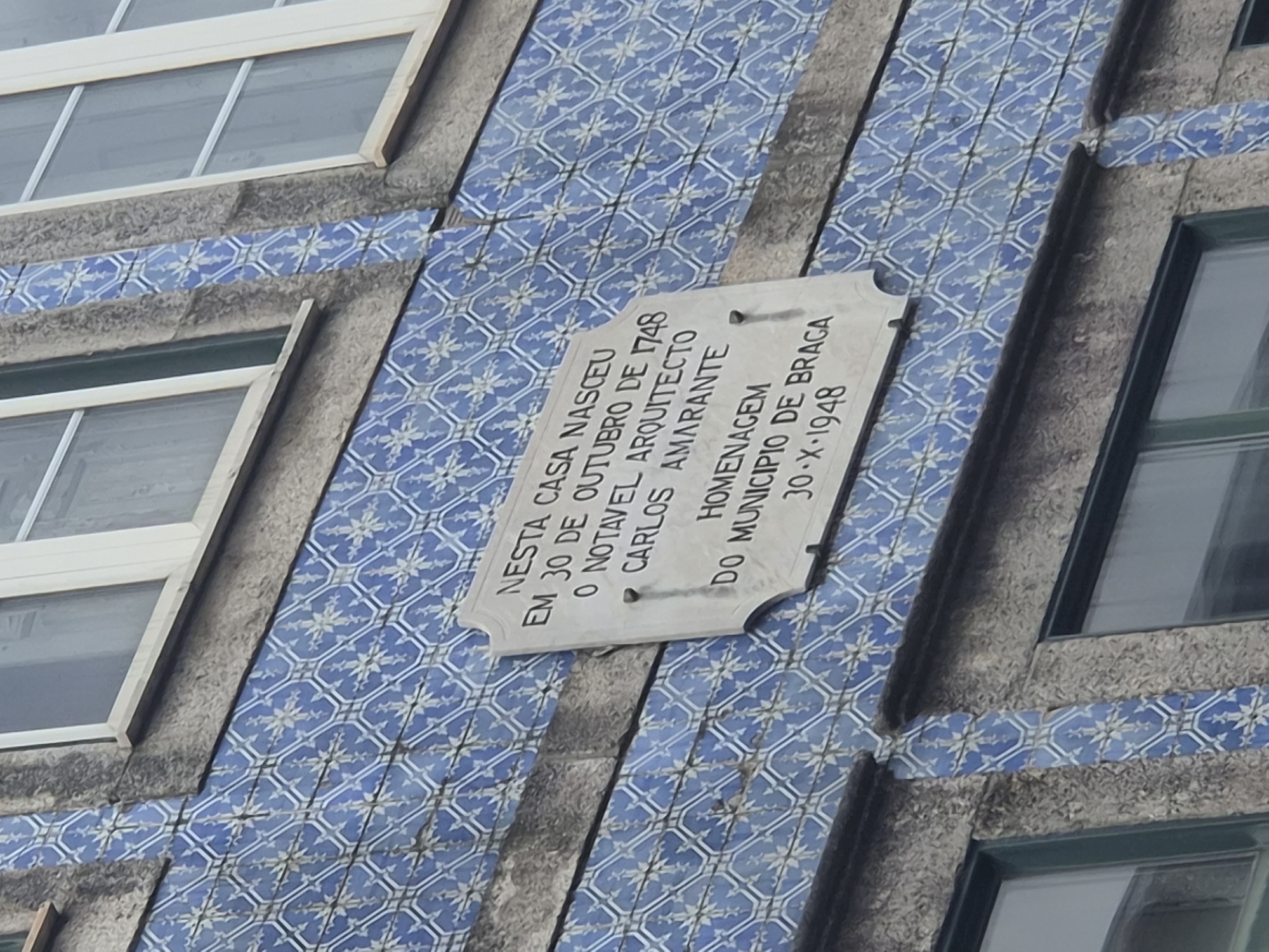 The image shows a close-up of a building facade in Braga, Portugal, decorated with traditional blue and white azulejo tiles. A white stone plaque is affixed to the tiled wall, bearing an inscription in Portuguese. The inscription reads: "NESTA CASA NASCEU EM 30 DE OUTUBRO DE 1748 O NOTAVEL ARQUITECTO CARLOS AMARANTE HOMENAGEM DO MUNICIPIO DE BRAGA 30-X-1948". This translates to: "IN THIS HOUSE WAS BORN ON OCTOBER 30, 1748 THE NOTABLE ARCHITECT CARLOS AMARANTE HOMAGE FROM THE MUNICIPALITY OF BRAGA 30-X-1948". The plaque commemorates the birth of Carlos Amarante and was erected by the municipality of Braga in 1948. The building features windows with dark frames. The overall impression is one of historical commemoration set against an aesthetically rich architectural background.