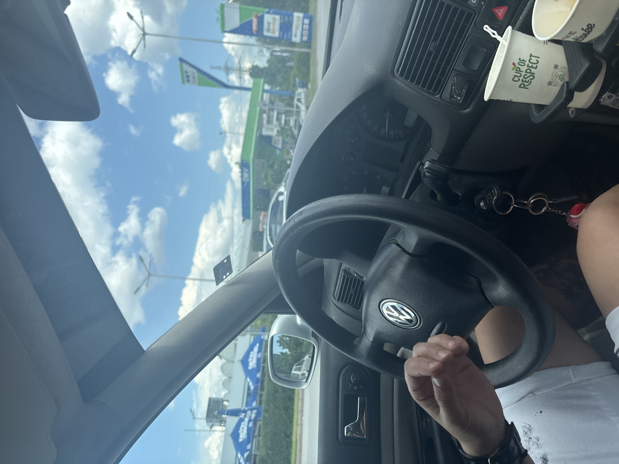 The image captures a first-person perspective from inside a car, likely a Volkswagen Golf (Mk4 generation), looking forward and slightly right. The driver's right arm and hand, wearing a watch, rest on the steering wheel, while their legs, clad in white distressed shorts, are visible below. Dangling keys with charms are attached to the ignition, and the dashboard with visible instruments and air vents is in view. In the lower right corner, two paper cups are placed in a cup holder; one is a "Cup of Respect" cup with a QR code, and the other is partially empty.
Outside the car, under a bright blue sky with scattered white clouds, a bustling commercial area is visible. To the left, through the side window and windshield, a main road runs alongside a green hedge, behind which are buildings with distinctive blue awnings and umbrellas, clearly marked with the Cyrillic text "Практикер" (Praktiker), indicating a large home improvement store.
Straight ahead and to the right, an OMV gas station is prominent. It features a large green canopy over the fuel pumps and a tall, vertical OMV price sign displaying various fuel options and prices. Beyond the gas station, a power line structure and other commercial or residential buildings are visible in the distance. The setting suggests a sunny day.
This scene is located in the city of Sofia, Bulgaria, specifically at the OMV gas station situated on Okolovrasten pat 216 (Sofia Ring Road), adjacent to a Praktiker store in the Mladost district.