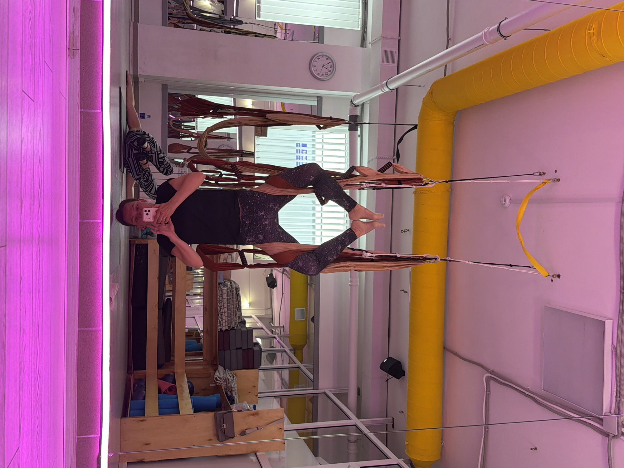A person is performing aerial yoga in a studio, suspended upside down in a yoga hammock. The person is wearing a black top and patterned leggings. Their feet are elevated, and they are holding a smartphone to take a photo of themselves. The studio is equipped with multiple yoga hammocks hanging from the ceiling, along with shelves stocked with yoga mats and other equipment. A large yellow pipe runs along the ceiling, and the room is lit with pink and white lights. A clock is visible on the wall in the background. The scene is set in a yoga studio in Chaikovsky, Russia.