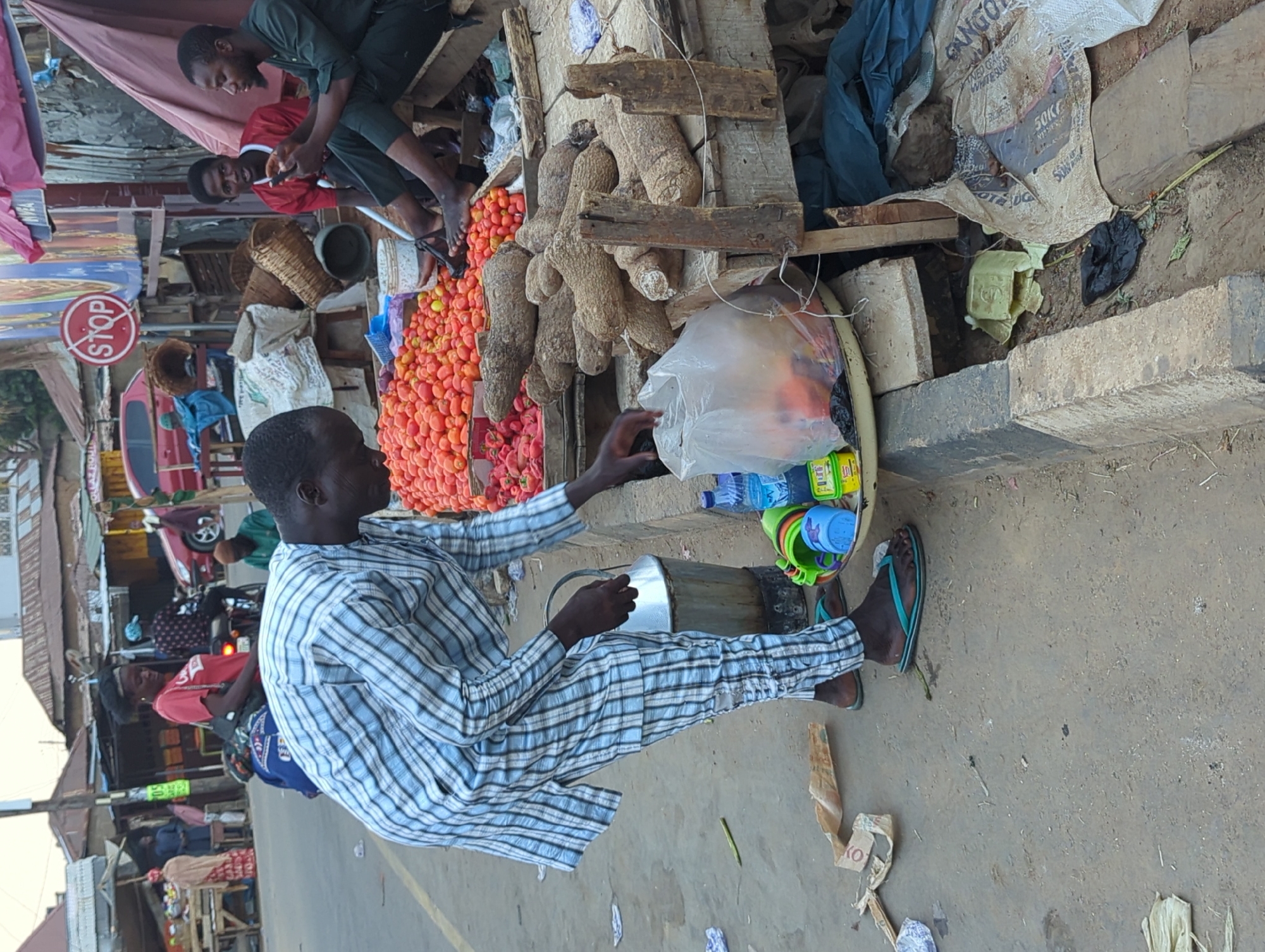 The image depicts a street scene in Shaba, Nigeria. A man in a blue and white striped shirt and pants sits on the ground next to a tray. The tray contains water bottles, small plastic cups, and a clear plastic bag with unknown contents. Next to the tray is an assortment of yams and tomatoes. Behind the tomato stand, two men are sitting and looking at a cell phone. A “STOP” sign is mounted nearby. In the background, other people are visible along the street with motorcycles and cars parked. The environment appears to be an open-air market area on a dirt and concrete surface.