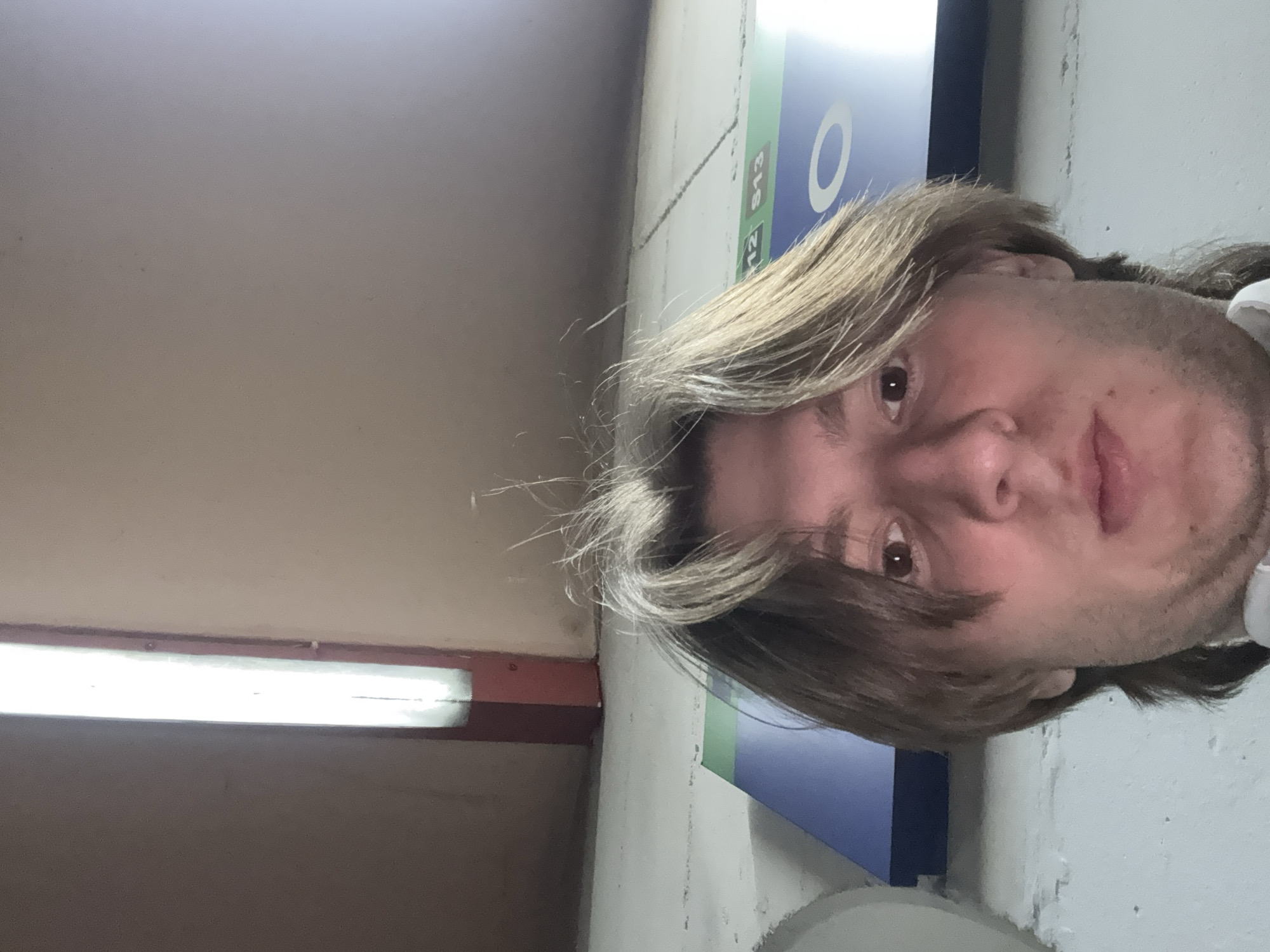 The image is a vertically oriented selfie, rotated 90 degrees clockwise, featuring a close-up of a person's face. The individual has medium-length, light brown to blonde hair, and brown eyes, looking towards the left side of the frame with a neutral expression. They have visible stubble.
The setting appears to be an indoor public space, possibly a transport station or a corridor. On the left side of the frame, a bright, vertically-running fluorescent light fixture is visible, set within a reddish-brown border. The walls are light-colored, likely beige or light grey, displaying some textured surface and minor scuffs.
At the top of the image, a blue sign with white text is partially visible. The discernible text reads "O", "S12", and "S13". Given the context of Milan, Italy, "S12" and "S13" likely refer to suburban railway lines within the Milan S Lines network. The artificial lighting suggests an indoor environment. No specific activity is taking place other than the person posing for the photo.