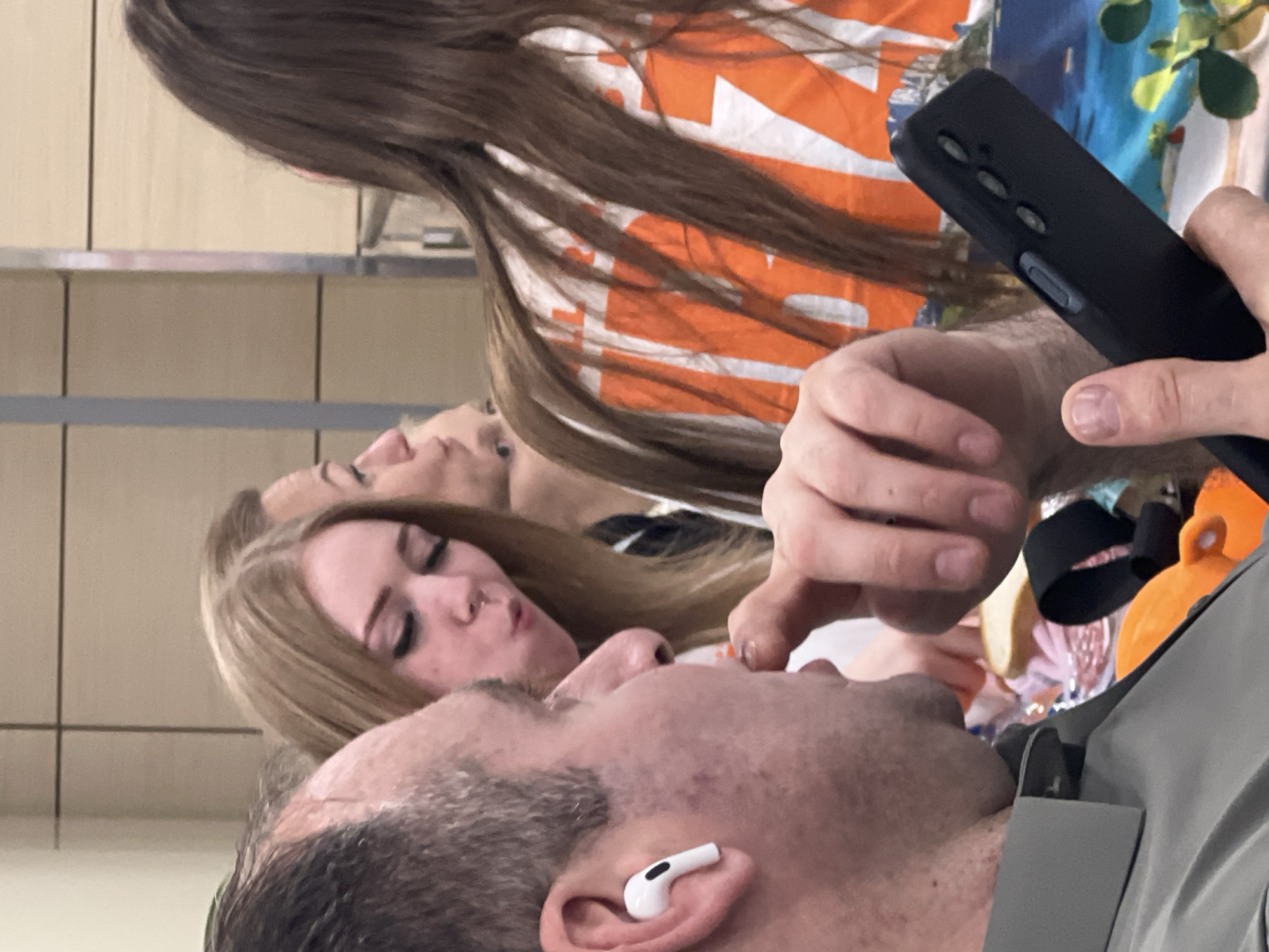 In Domodedovo, Russia, a close-up shot captures a man in the foreground wearing a grey shirt and a white earbud in his left ear. He is holding a black smartphone in his right hand, with his fingers obscuring part of the device. His right thumb is extended towards his mouth. Behind him, a young woman with blonde hair and visible nose jewelry is looking downwards. Further back, another individual's face and long brown hair are visible, along with part of an orange and white patterned shirt. The background appears to be an indoor setting with light-colored walls and a vertical divider, possibly a public space like an airport or train station.