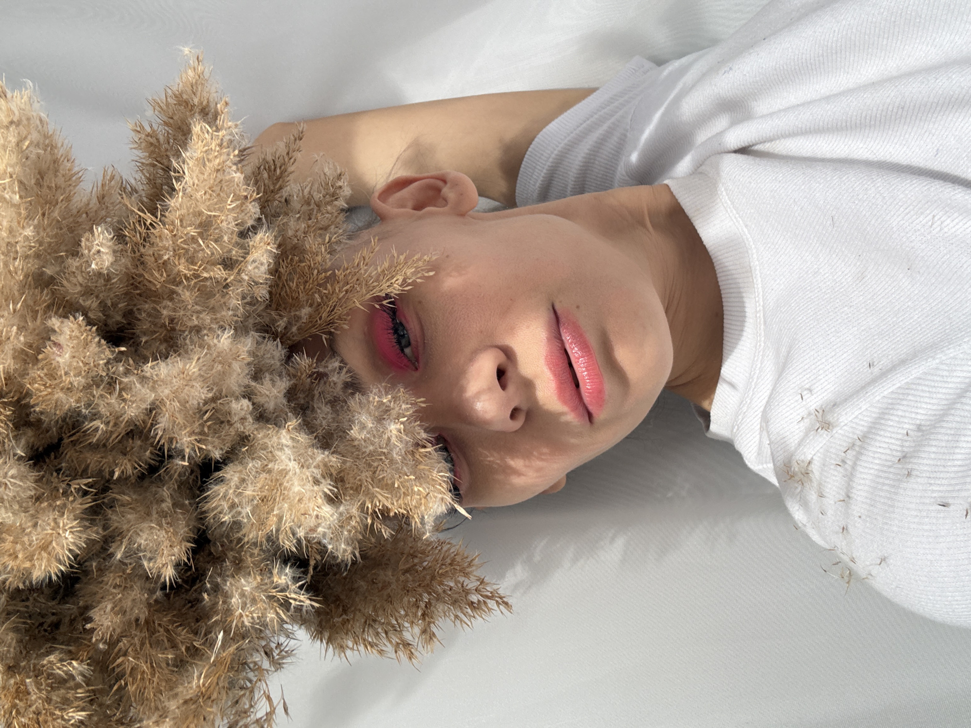 A woman is lying on a white surface, presumably in an indoor location. She is wearing a white top and has pink makeup around her eyes and on her lips. A cluster of pampas grass is positioned next to her head, some of the feathery plumes touching her face. There are seeds scattered on her clothing. The lighting suggests an indoor environment with natural light sources creating shadows. Location cannot be determined from the image.