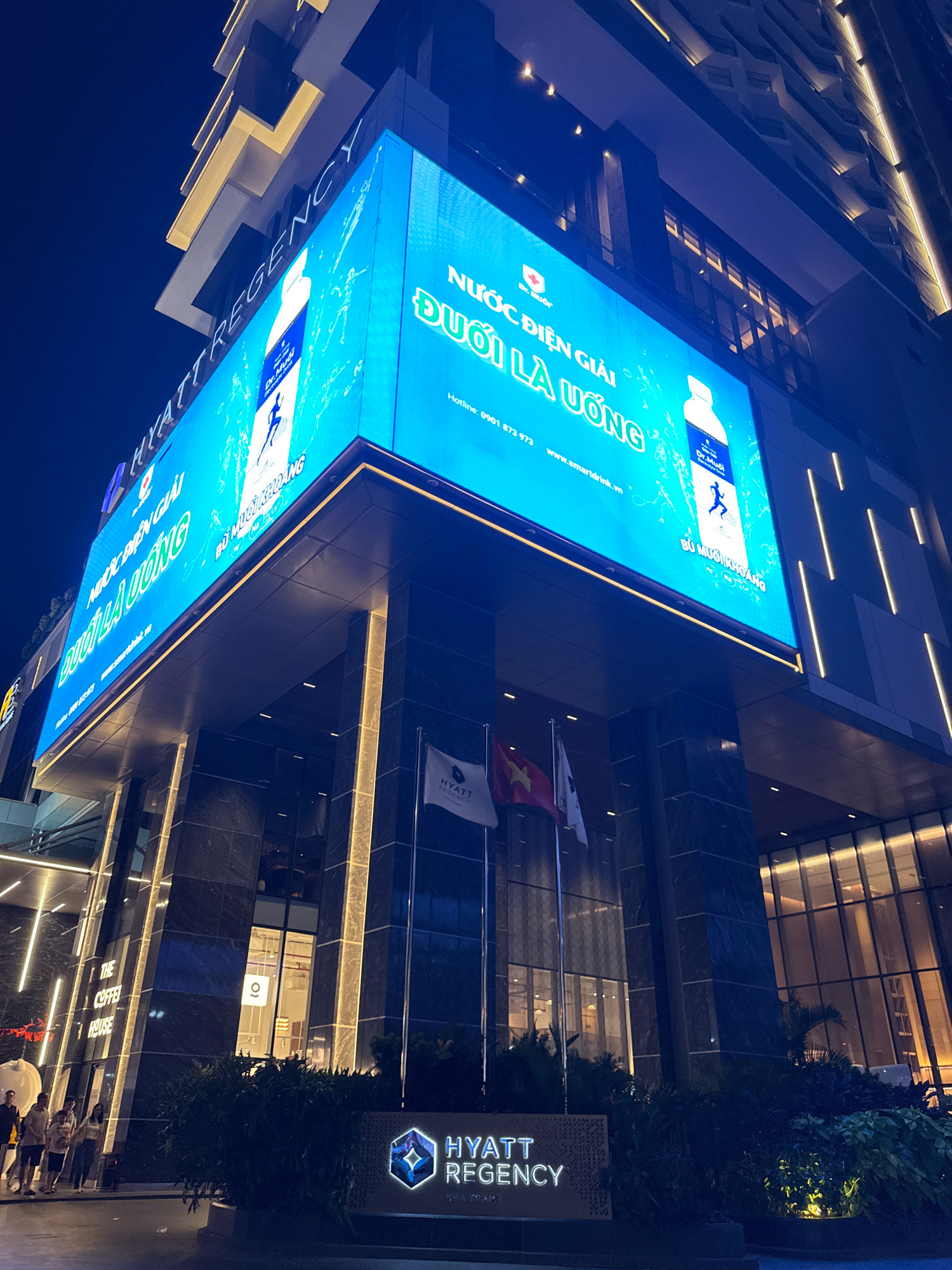 The image depicts the Hyatt Regency Nha Trang hotel entrance at night. A large LED screen is prominently displayed, advertising "Dr. Muối" electrolyte drink, featuring text in Vietnamese and images of a water bottle and a running figure. The hotel's name, "HYATT REGENCY," is visible in large letters above the screen and also as a lit sign near the bottom of the image. Flags are planted near the entrance, one of which is the Vietnamese flag. Several people are walking near the entrance, some appearing to be hotel guests. The architecture of the hotel is modern, with illuminated vertical lines on the facade. The scene is set against a dark night sky, with the building's lights illuminating the area. The overall impression is one of a well-lit, upscale hotel entrance in a city setting.