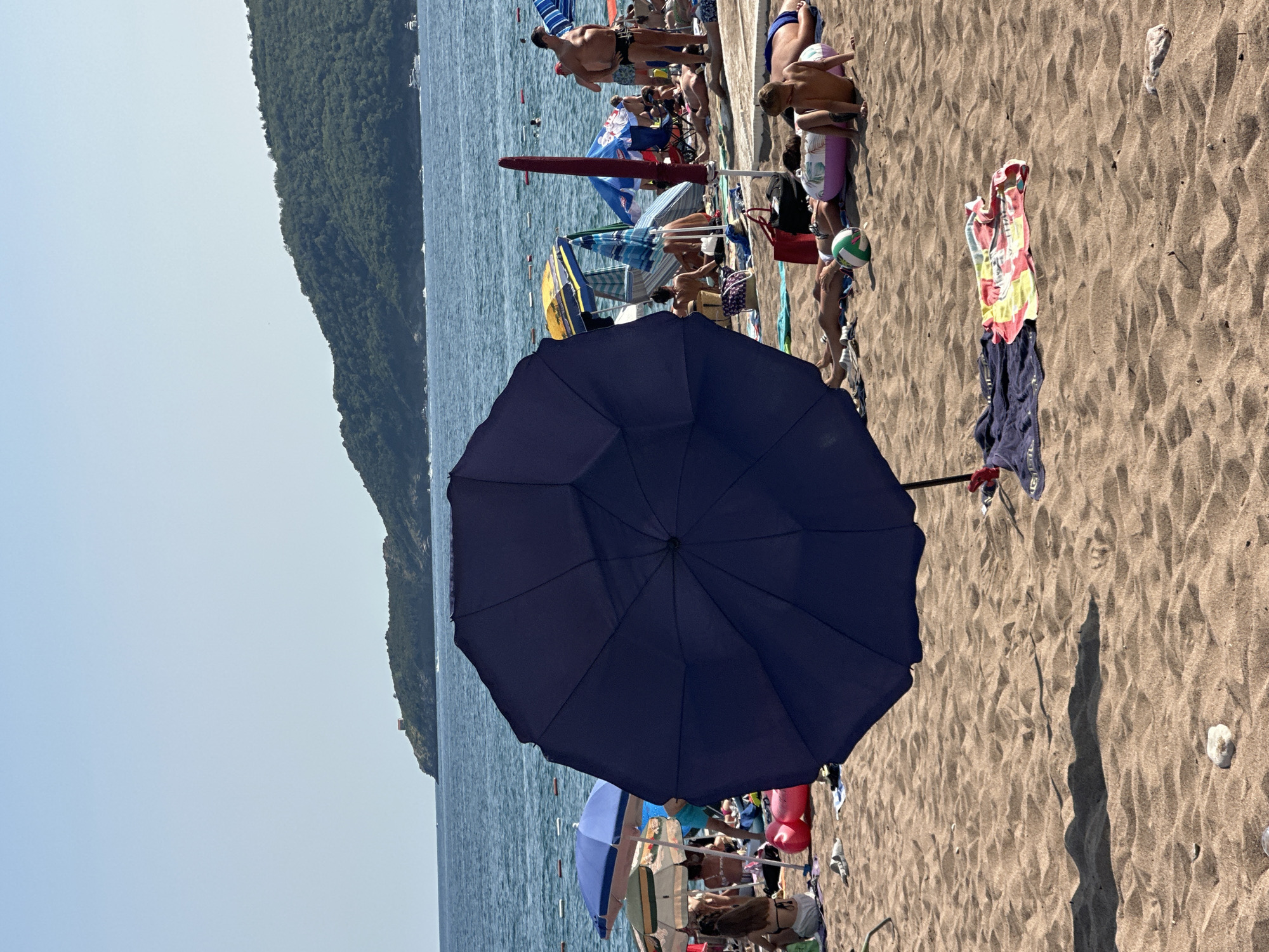 This image captures a sunny day on a crowded beach in Boreti, Montenegro. The foreground is dominated by a large, dark blue umbrella, casting a shadow across the sand. Scattered on the sand are various beach accessories, including towels, a striped beach mat, a green and white ball, and a pink inflatable ring. A few people are visible in the background, either lying on towels or sitting under other umbrellas. The sea stretches out to the horizon, with a hilly, tree-covered coastline visible in the distance. Several boats are scattered across the water, and a few people are swimming. The overall scene depicts a typical summer day at a popular beach destination.