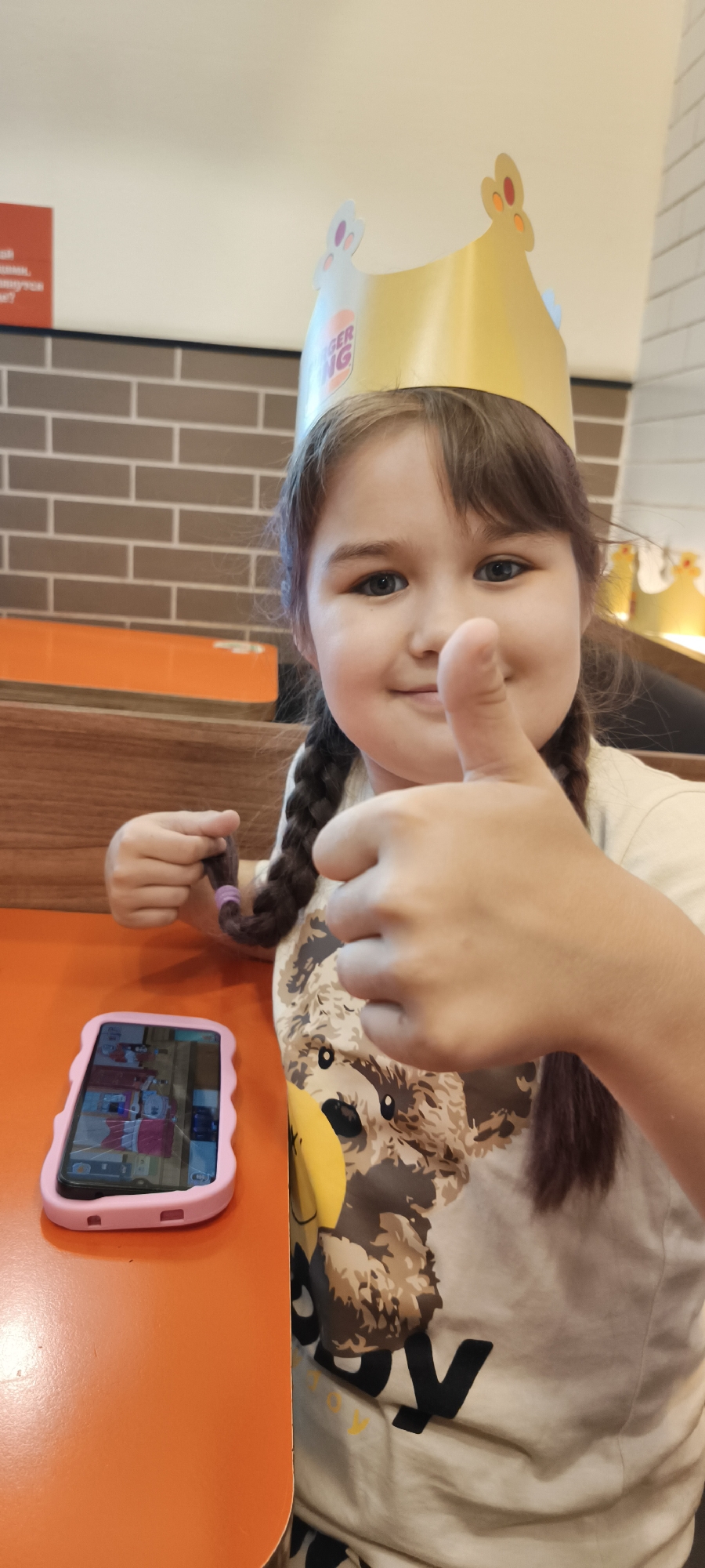 A young girl wearing a golden Burger King crown and a t-shirt with a teddy bear graphic is shown in a close-up shot, giving a thumbs-up. Her hair is in two braids, and she is holding one of her braids with her left hand. A pink-cased smartphone is visible on an orange table in front of her, displaying what appears to be a game or app. The background shows a brick-patterned wall and what looks like a Burger King restaurant interior, with orange and wooden furniture. The girl appears happy and engaged. The image is well-lit, suggesting it was taken indoors during the daytime. The text "BURGER KING" is visible on the crown.