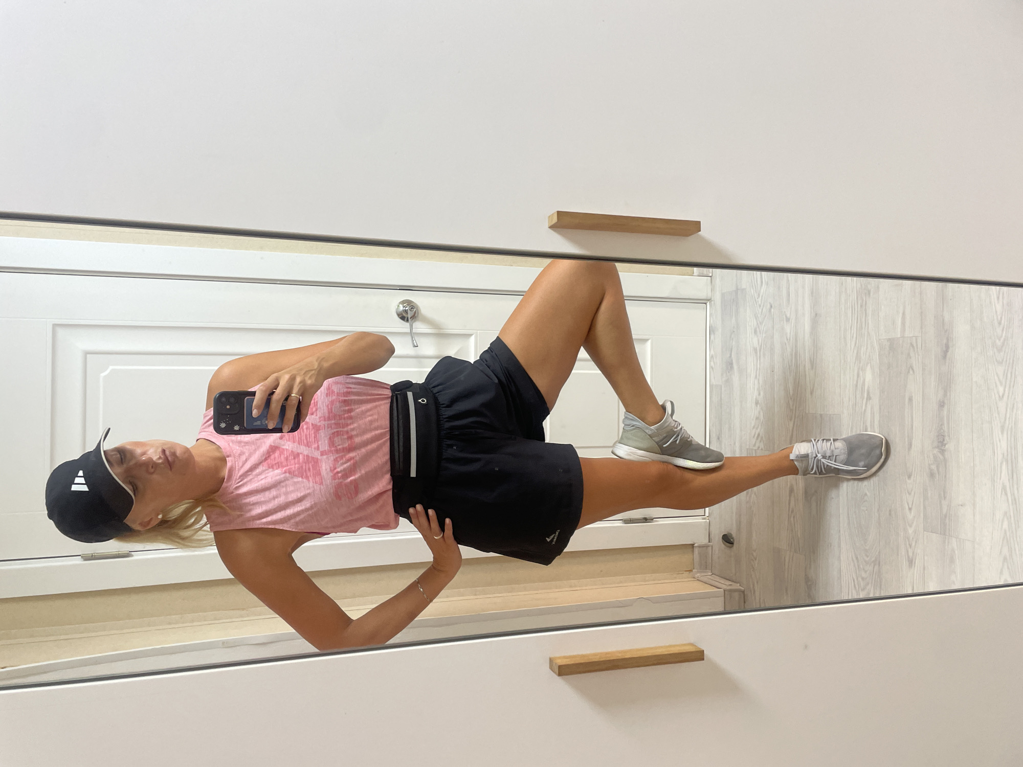 A woman wearing a black baseball cap, a pink sleeveless top, black athletic shorts, and grey athletic shoes is standing in front of a full-length mirror. She is holding a smartphone in her right hand, taking a selfie. Her left hand is on her hip. Her right leg is bent at the knee and resting on the mirrored surface, with her foot lifted. The background shows a white door and a light-colored wooden floor. There is no visible indication of the city of Baranovka, Russia.