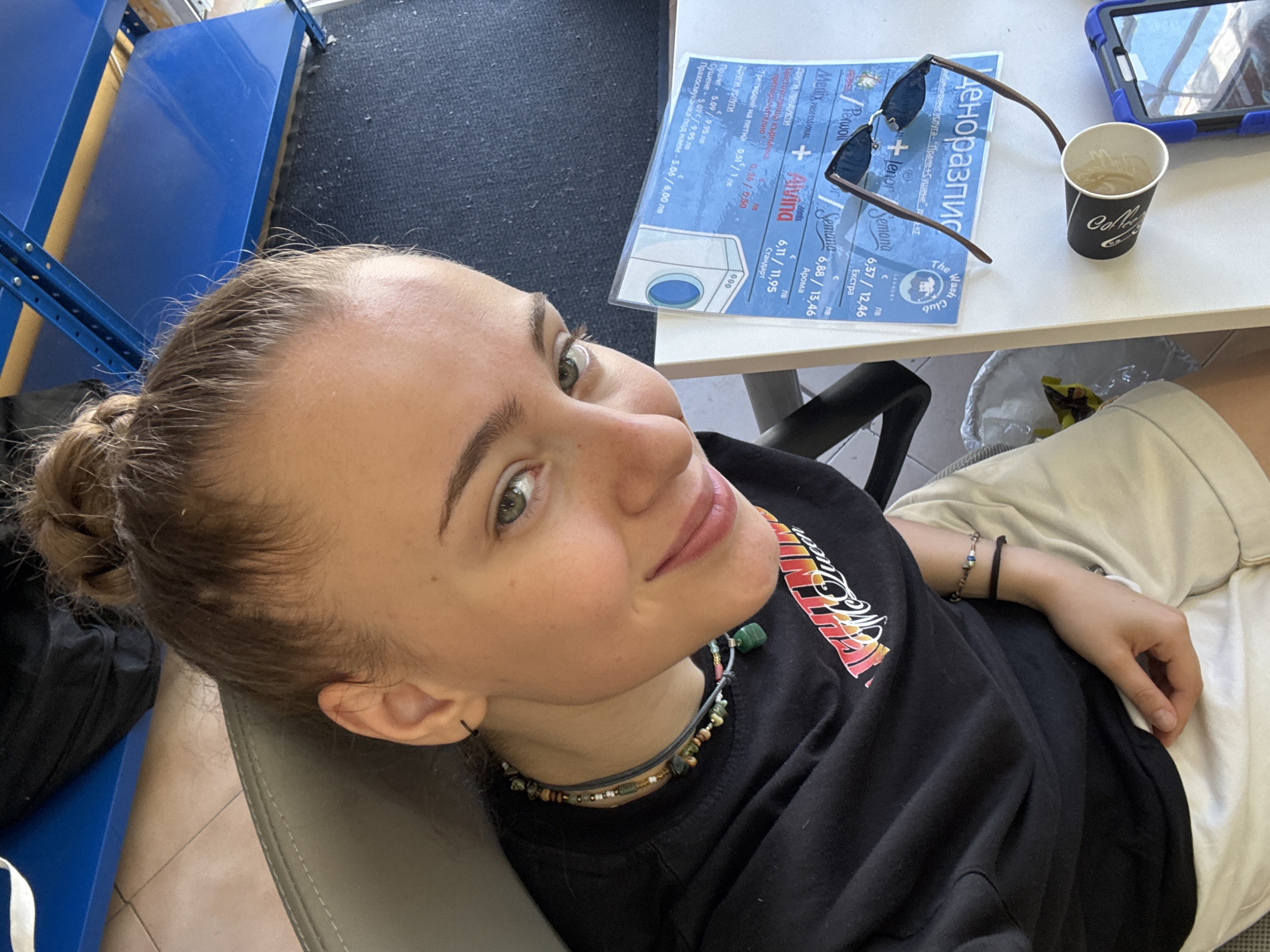 A young woman with her hair in a bun, wearing a black t-shirt with a graphic print and light-colored shorts, smiles at the camera. She is seated at a table with a blue and white sign, sunglasses, and a paper cup. The scene appears to be indoors, possibly a cafe or a laundromat, with blue shelving visible in the background. The table has some text in Cyrillic script. The overall lighting is bright.
