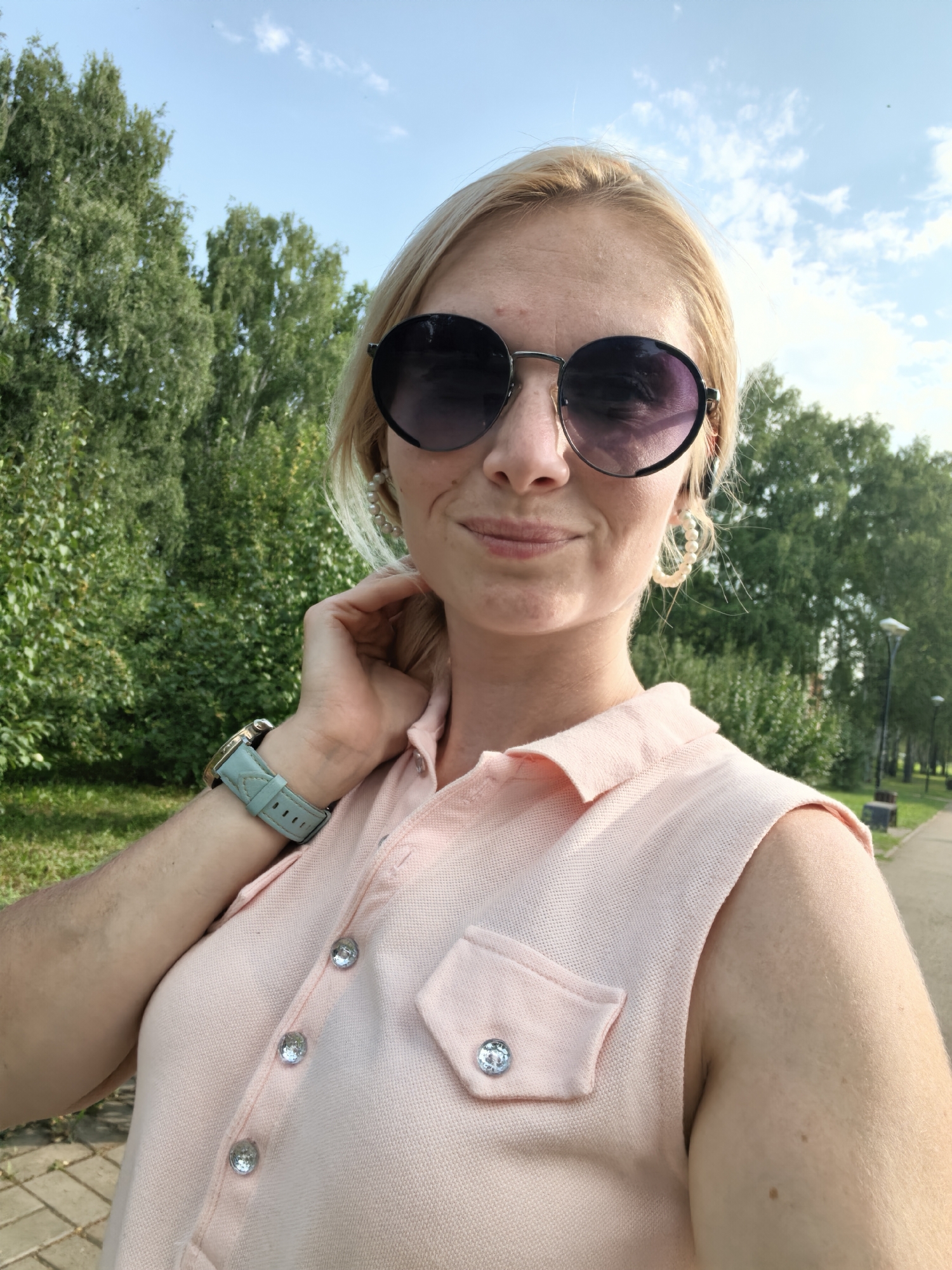 A woman with blonde hair and sunglasses takes a selfie outdoors on a paved path. She is wearing a pink sleeveless polo shirt with sparkling buttons and a light blue watch on her left wrist. She has pearl hoop earrings. The background shows lush green trees and a clear blue sky with scattered clouds. The scene appears to be a park or a tree-lined street.