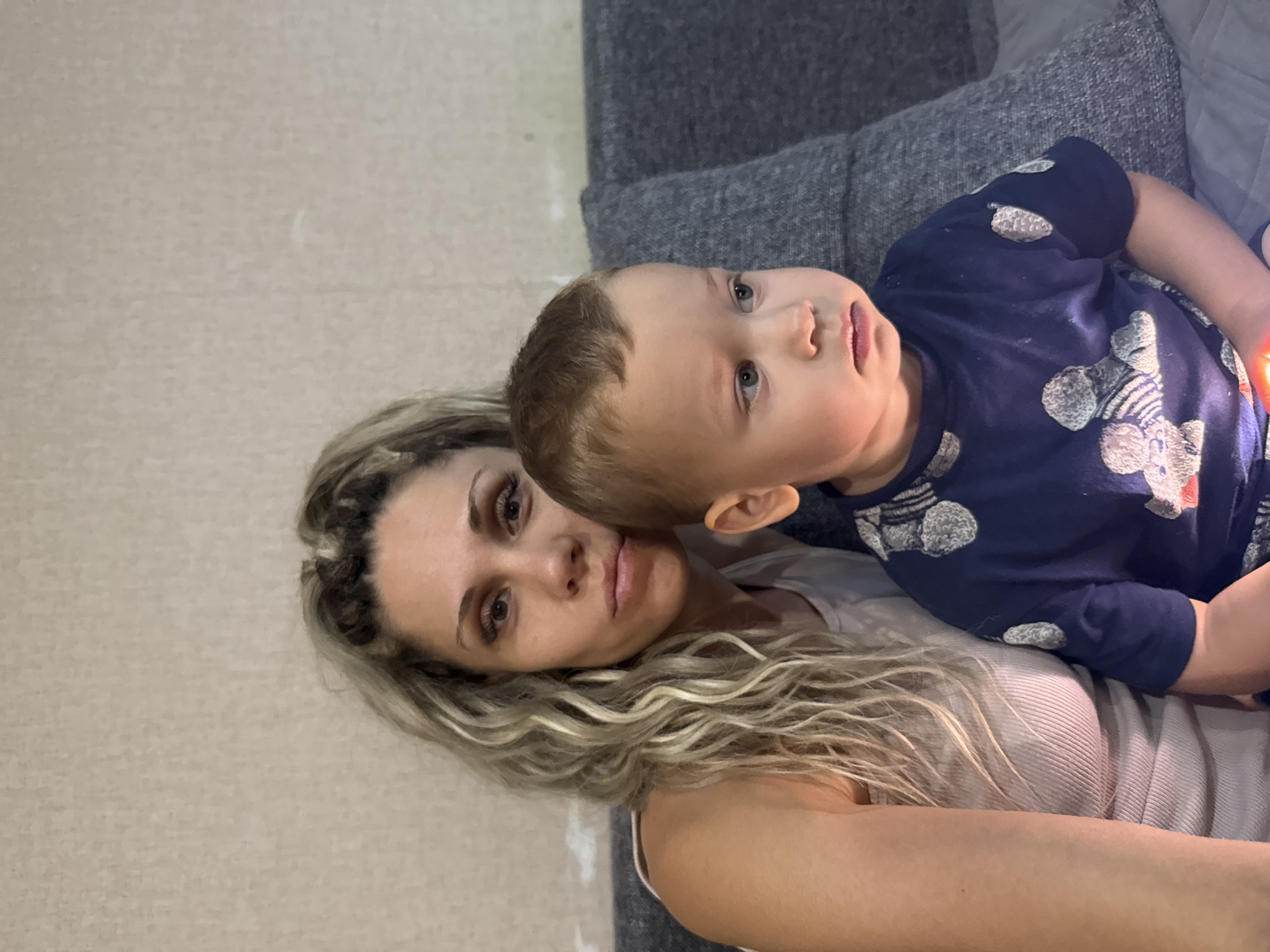 An indoor, medium shot shows a woman and a young boy posing for a selfie against a textured, light-colored wall and a grey sofa. The woman, with long, curly blonde hair, is on the left, wearing a light-colored, ribbed tank top. She has a neutral expression and is looking towards the camera. The boy, on the right, has short brown hair and is wearing a dark blue t-shirt with a pattern of grey and white teddy bears. He is looking to the right with a pensive expression. The lighting suggests an indoor environment. The location is identified as Rusanovo, Russia.