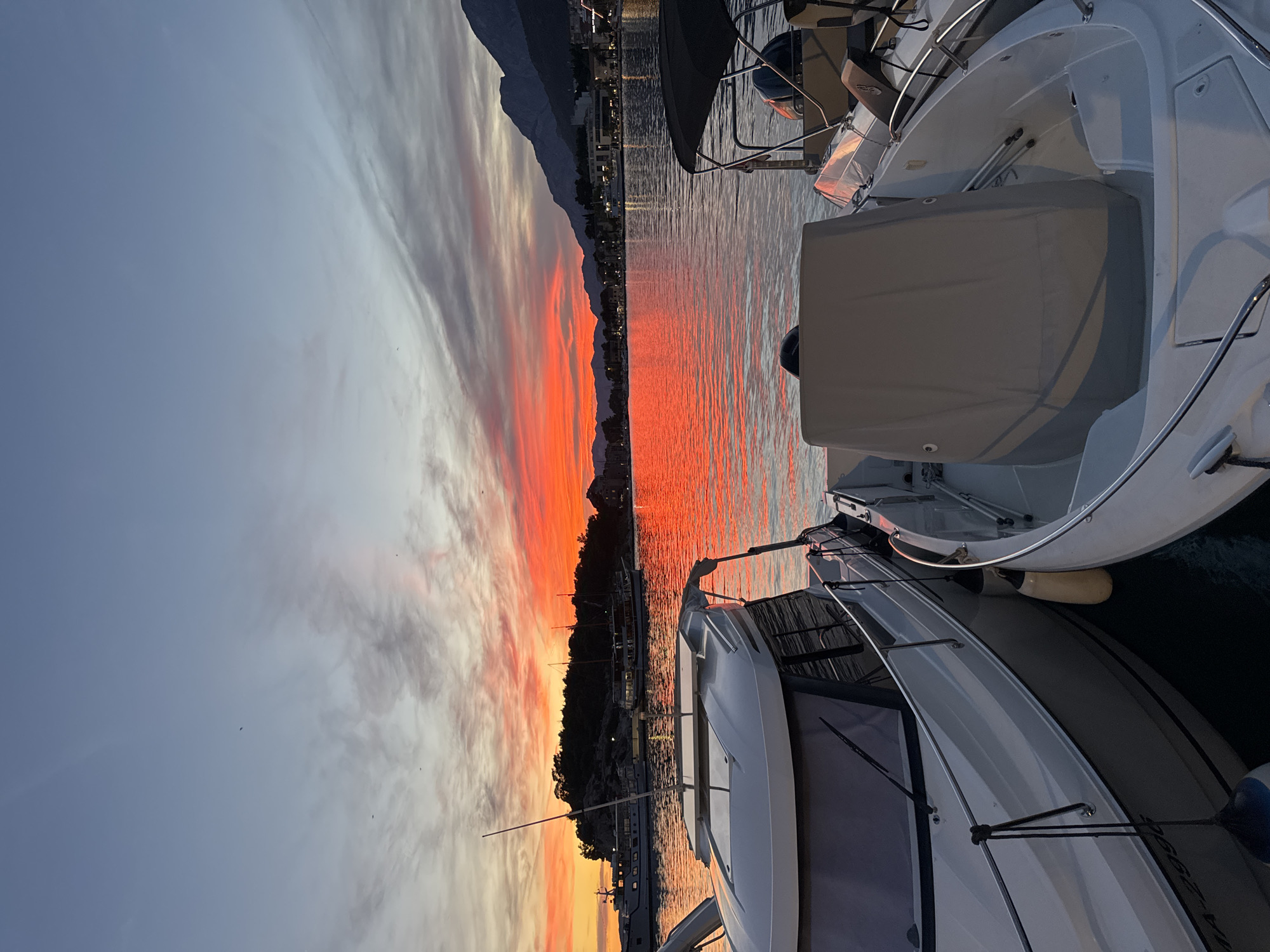 A vibrant sunset casts a fiery orange glow across the water and sky in Makarska, Croatia. The scene is dominated by several boats docked in the harbor, with their white hulls reflecting the dramatic colors of the twilight. In the foreground, two boats are prominently featured, their sleek designs indicative of leisure craft. The boat on the left is larger and more enclosed, while the one on the right is smaller and more open, with a canopy.
Beyond the boats, the calm water mirrors the intense hues of the sunset, creating a liquid inferno of reds and oranges. In the distance, a tree-covered island with a rocky silhouette sits on the left, and a coastal town with buildings and lights stretches along the shoreline to the right. Majestic mountains rise behind the town, their peaks silhouetted against the colorful sky. The sky itself transitions from a soft blue at the top to fiery oranges and reds near the horizon, with wispy clouds catching the last rays of the sun. A few small birds are visible in the sky. The overall atmosphere is one of serene beauty and tranquility, typical of a Mediterranean coastal evening.