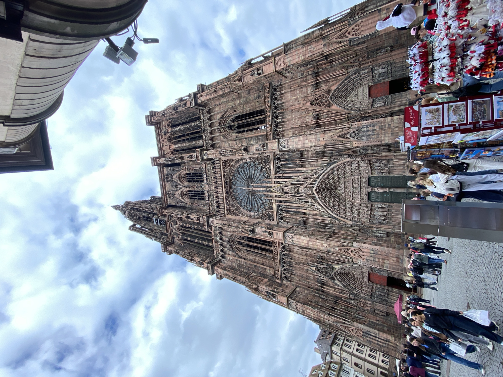 The image captures a view of the Cathedral Notre Dame de Strasbourg in Strasbourg, France. The cathedral's exterior facade is visible in detail, showcasing its Gothic architecture. A crowd of people is gathered in front of the cathedral, suggesting a popular tourist destination. A vendor stall selling souvenirs is also present near the entrance. The sky is overcast with clouds.