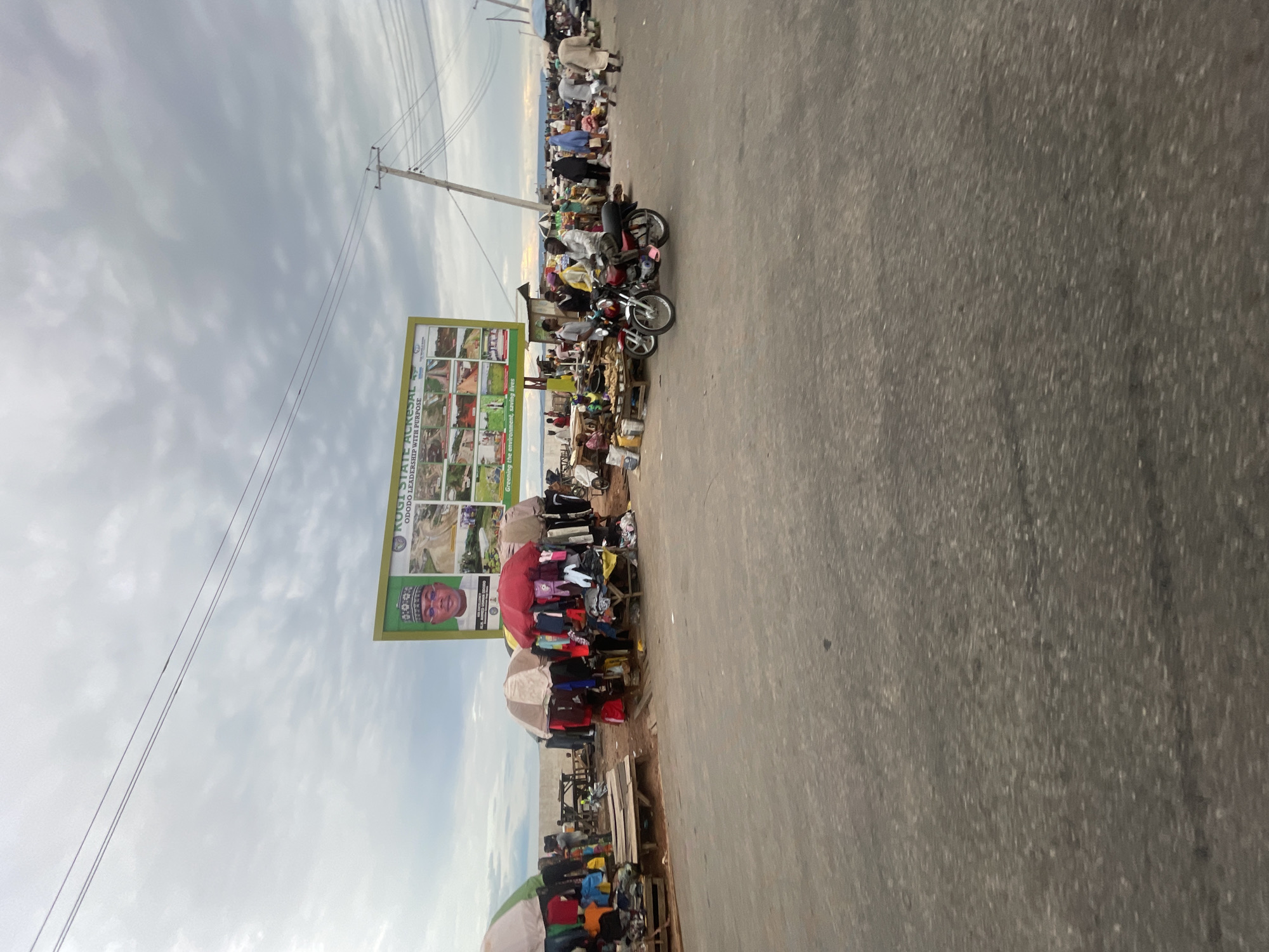 The image shows a street scene in Lokoja, Nigeria. On the left side of the image, there are numerous stalls selling goods, with people browsing and interacting with the vendors. There are umbrellas providing shade over the stalls. A billboard is visible on the left side of the image, featuring the image of a man wearing eyeglasses and a traditional cap. Motorbikes are parked near the stalls. Overhead, power lines run along the street. The sky is cloudy. The road occupies a large portion of the right side of the image.