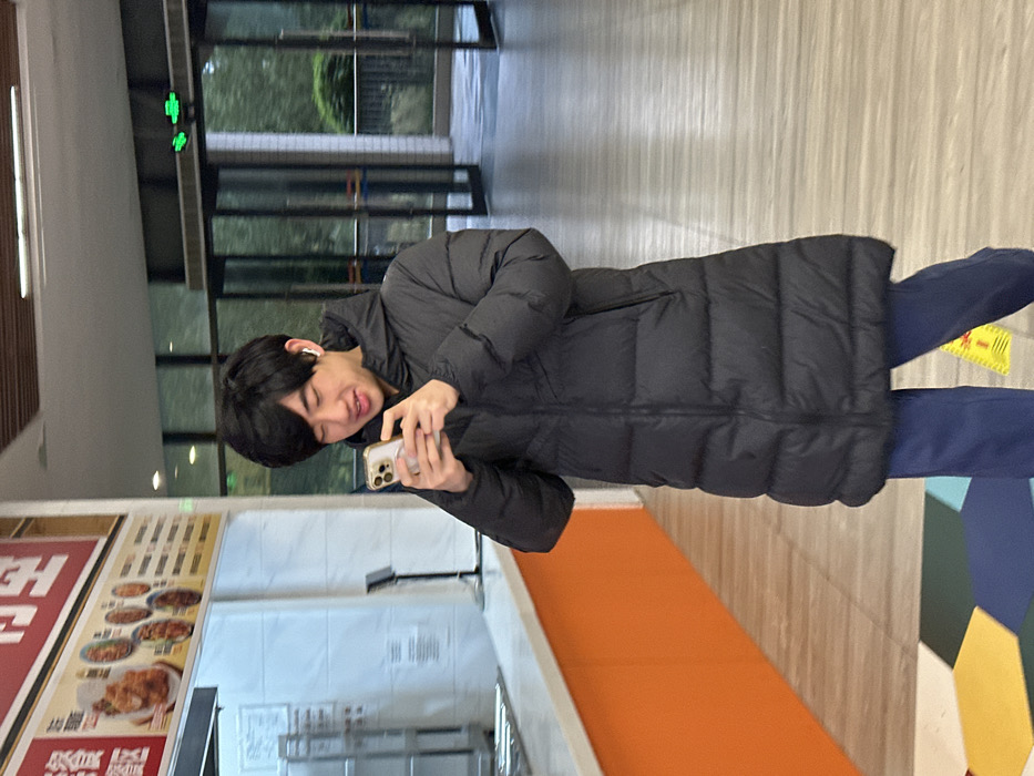 An East Asian male, appearing to be in his late teens or early twenties, stands centrally within a modern indoor cafeteria or dining facility in Jiangjin, China. He has short, dark hair and wears a black, hooded, long-length puffer coat, blue trousers, and white wireless earbuds in both ears. He is holding a light-colored smartphone horizontally with both hands, looking down at the screen with a slight smile and open mouth, actively engaged with the device.
To the left of the subject, a food service counter with an orange base is visible beneath a red signboard displaying white Chinese characters and a menu board with images of various dishes. Stainless steel kitchen equipment is partially visible behind the counter. To the right, large glass double doors, partially ajar, open to an outdoor area with visible greenery. A green exit sign is mounted above these doors. The floor is covered with light-colored tiles, with a section of multi-colored hexagonal tiles (dark blue, light blue, yellow, orange) in the foreground bottom left. A yellow warning sign with red text is partially visible on the floor near the subject's right foot. The area is illuminated by overhead interior lighting and natural light from the exterior.