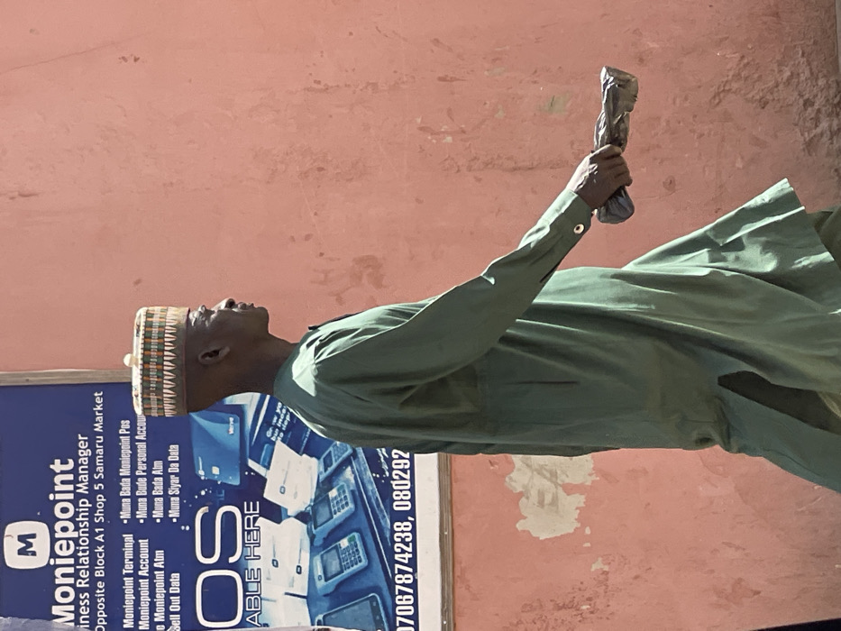 A male subject is captured in profile, walking from left to right, in an outdoor setting in Samaru, Nigeria. He wears a light olive-green long-sleeved tunic and matching trousers, topped with a round, patterned hat featuring a central knob. His right hand holds a dark, irregularly shaped object, possibly wrapped in black material. The background consists of a reddish-orange building wall exhibiting significant paint wear and discoloration. To the left, a large blue and white commercial sign for "Moniepoint Business Relationship Manager" is partially visible. The sign specifies the location as "Opposite Block A1 Shop 5 Samaru Market" and advertises services including "Moniepoint Terminal," "Moniepoint Account," and "Moniepoint ATM," displaying graphics of POS devices and a Verve card logo. The scene is brightly lit by natural sunlight.