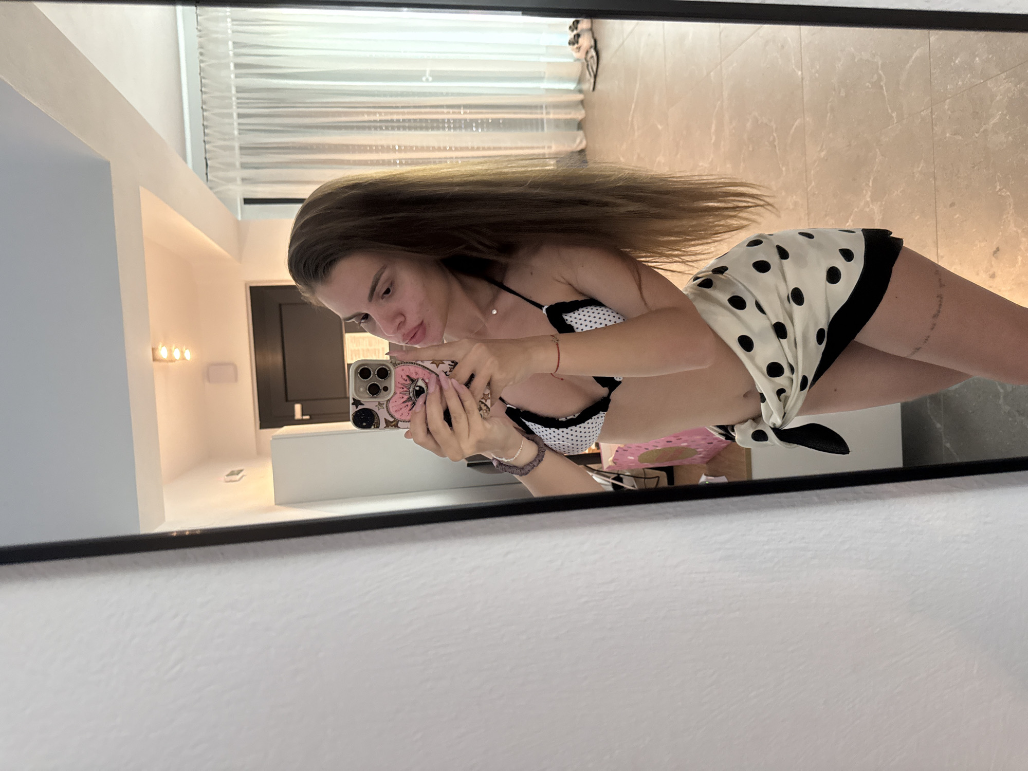 A mirror selfie captures a woman wearing a black and white polka dot bikini top, with a matching sarong tied at her waist, revealing her lower body. She's holding a smartphone with a decorated case, presumably taking the picture. The background reveals a well-lit interior, including a marble-like tiled floor, a window with blinds, a doorway, and a white cabinet. The scene appears to be within a residential space, though the specific location in Unknown, Unknown cannot be identified.